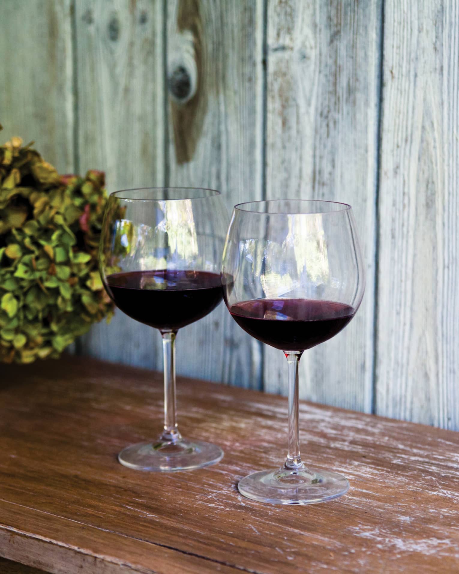 Two glasses of red wine and sit on wooden table, with white vase filled with hydrangeas
