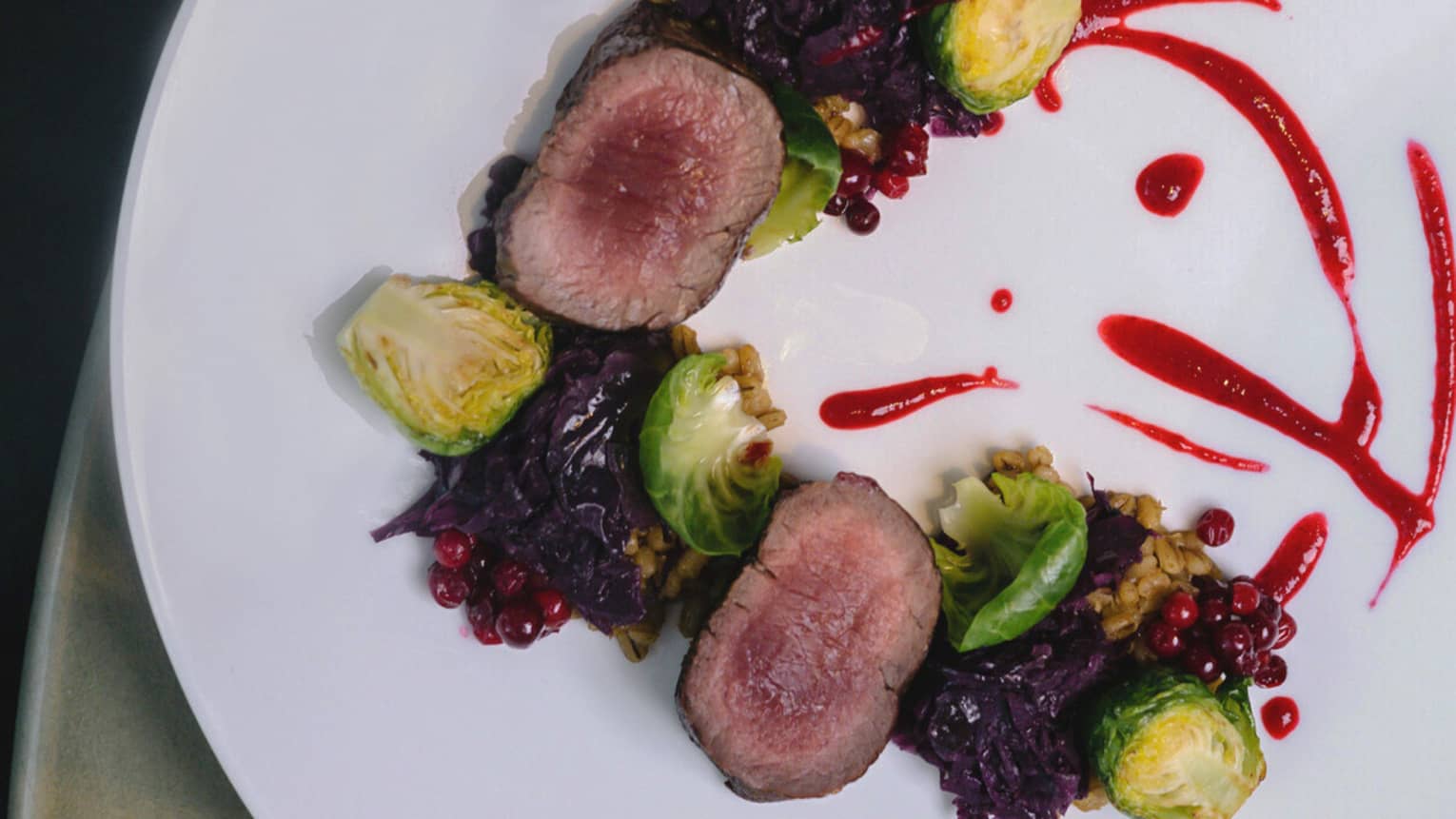 Two slices of venison, halved Brussels sprouts, red cabbage and lingonberries arranged in a half circle with streaks of red-colored sauce on the other side all on a round white plate