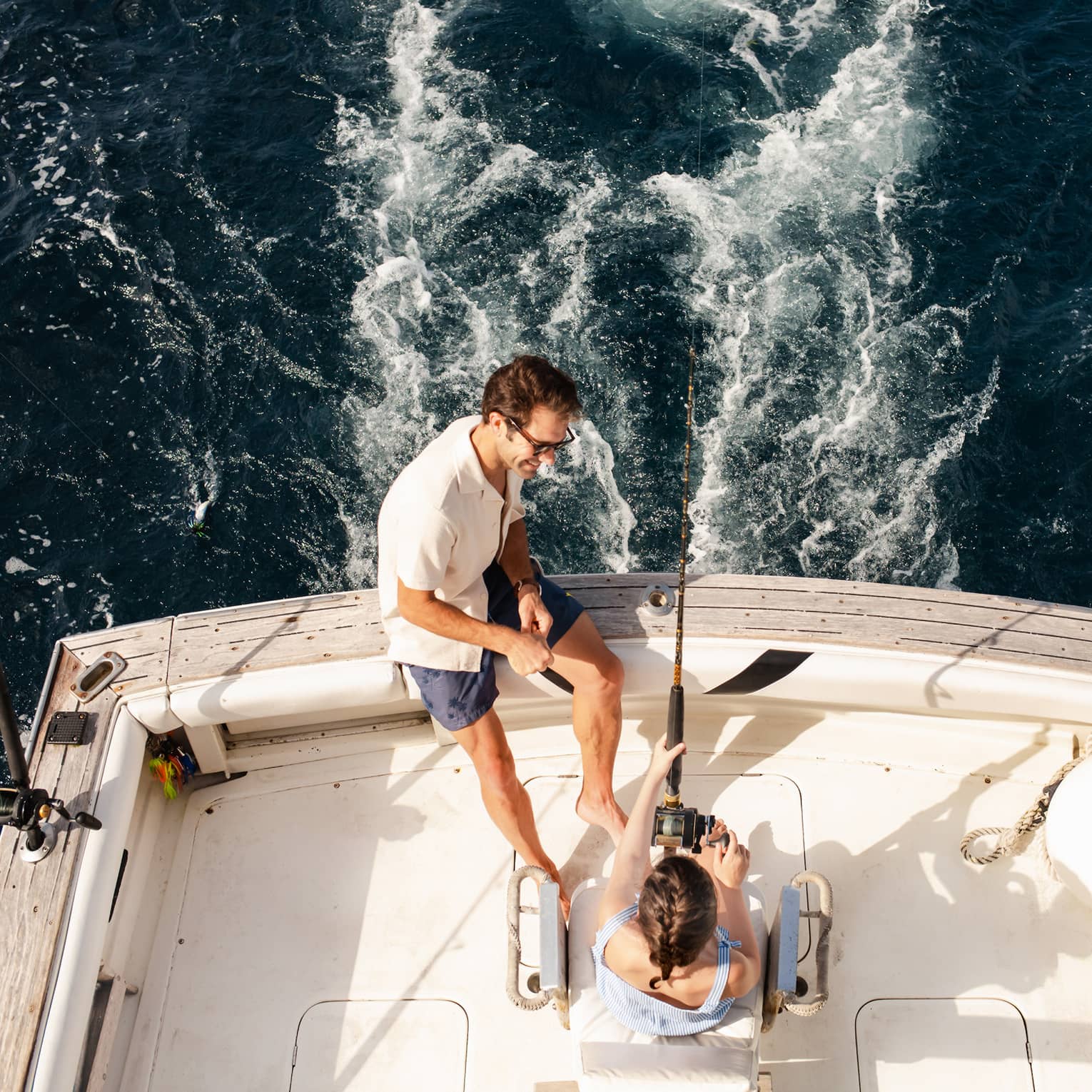 Two people sit at the back of a fishing boat, with one person holding a fishing rod and the other sitting close by on the edge of the boat