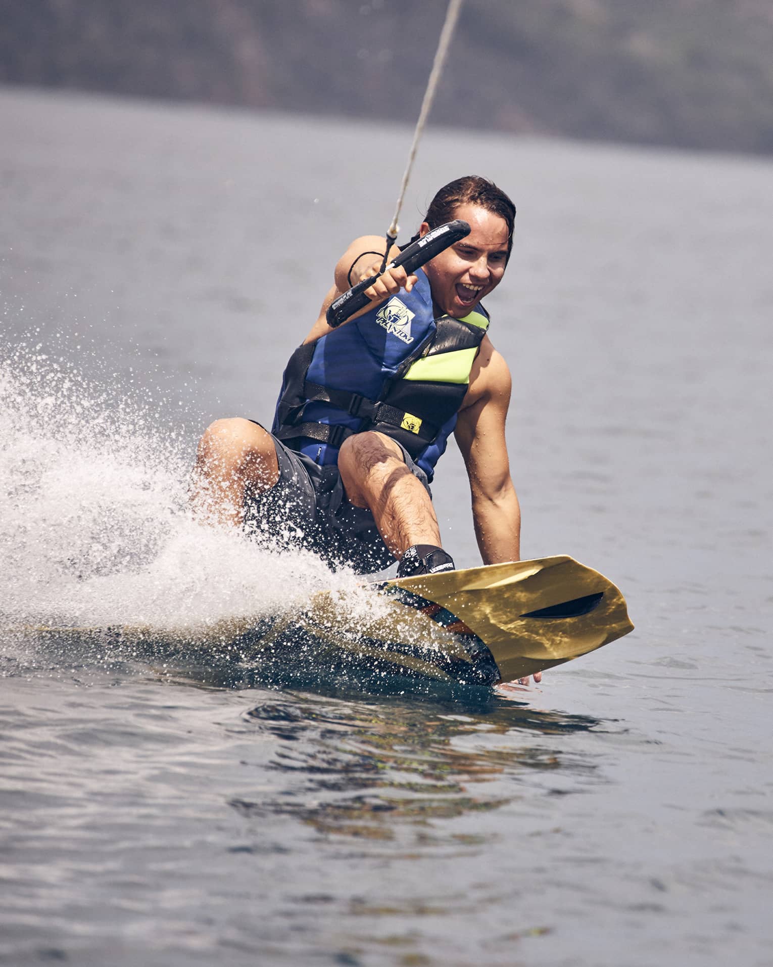 Young man wearing a blue and green life vest and blue swim trunks wakeboarding through the water