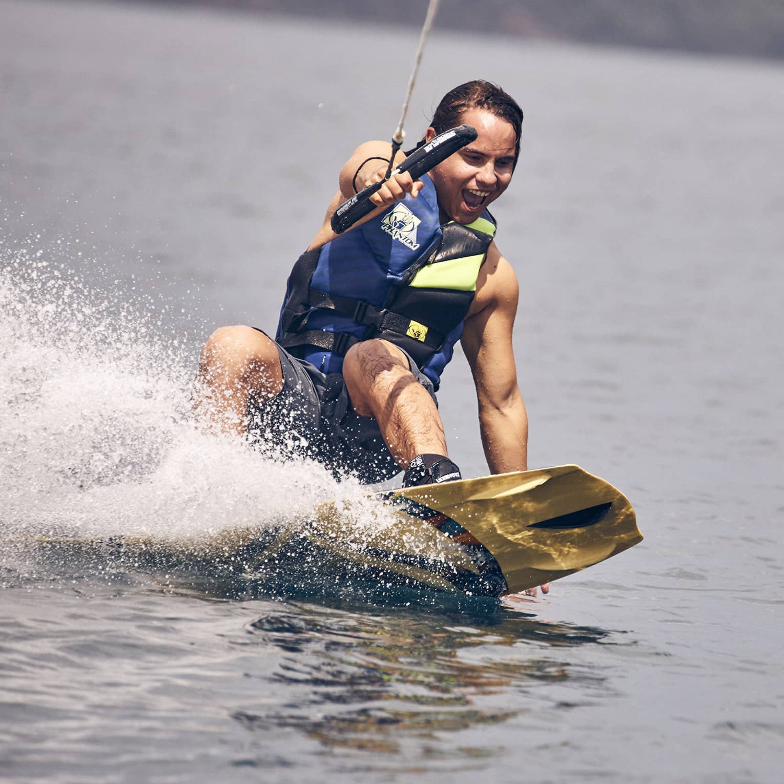 Young man wearing a blue and green life vest and blue swim trunks wakeboarding through the water