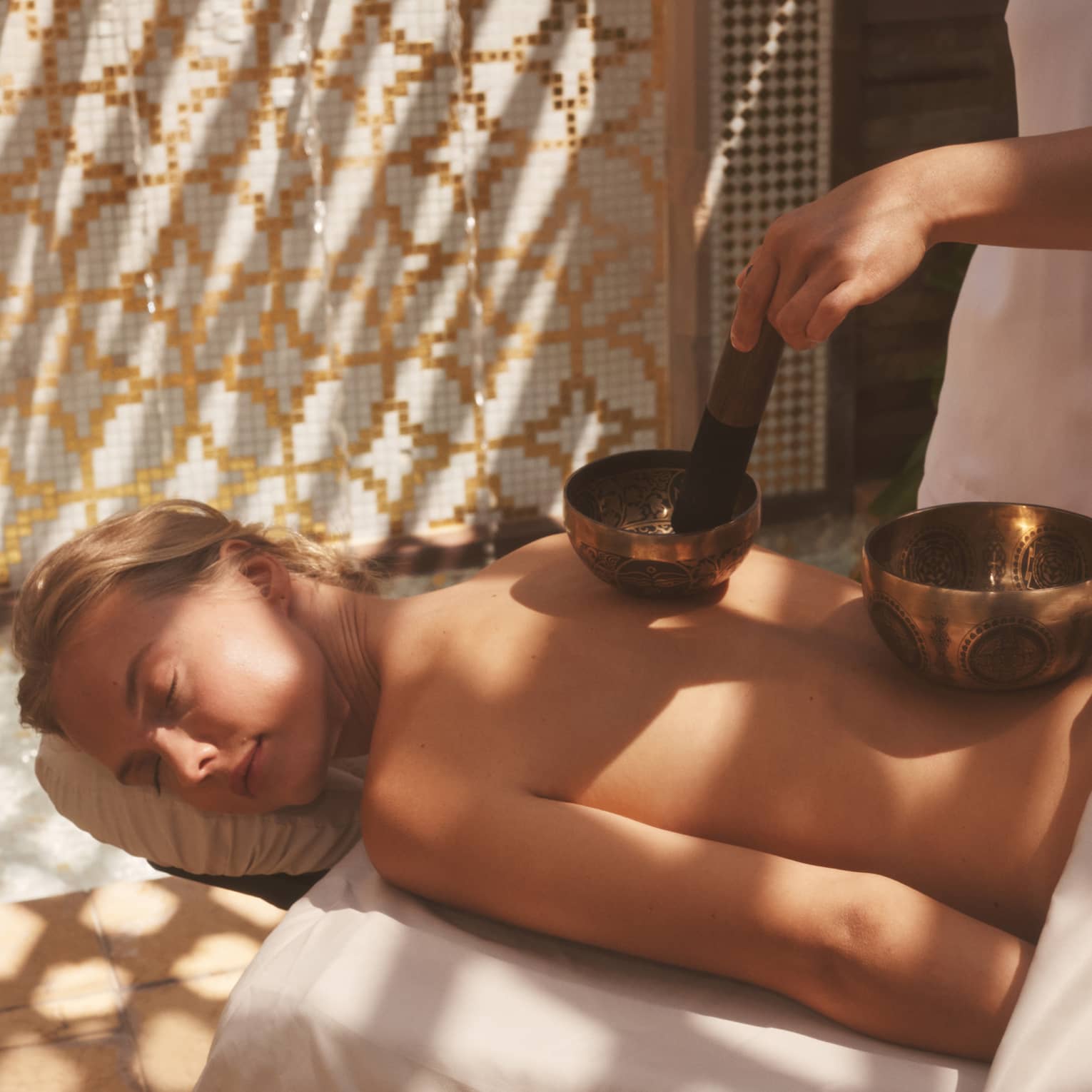 Unclothed person lying prone on a spa table with two sound healing bowls on back and spa therapist using them