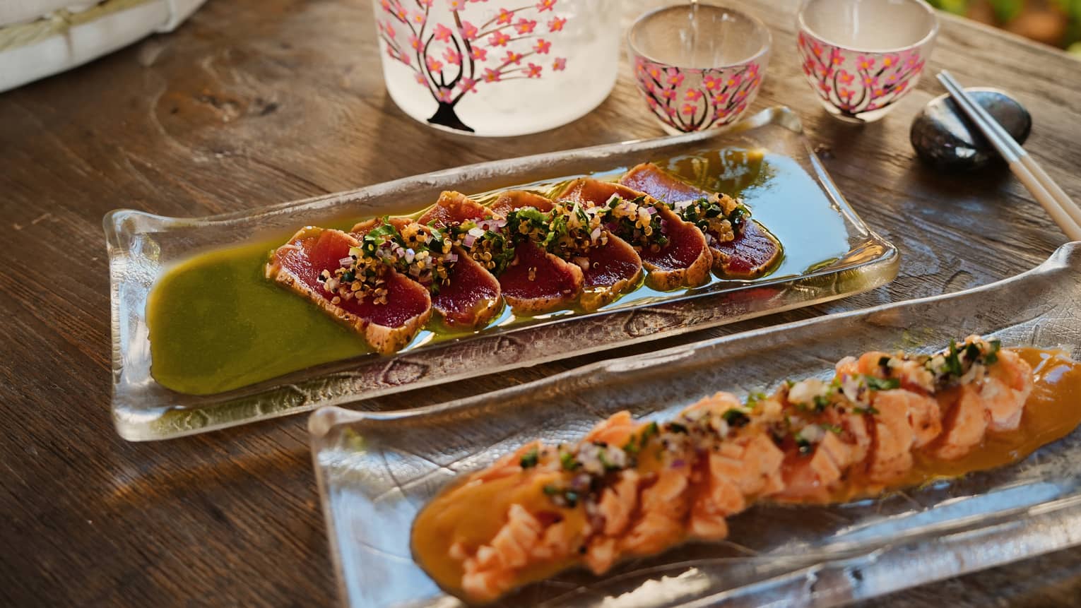 A person pours liquid into a small cup next to two rectangular plates of colourful food items.
