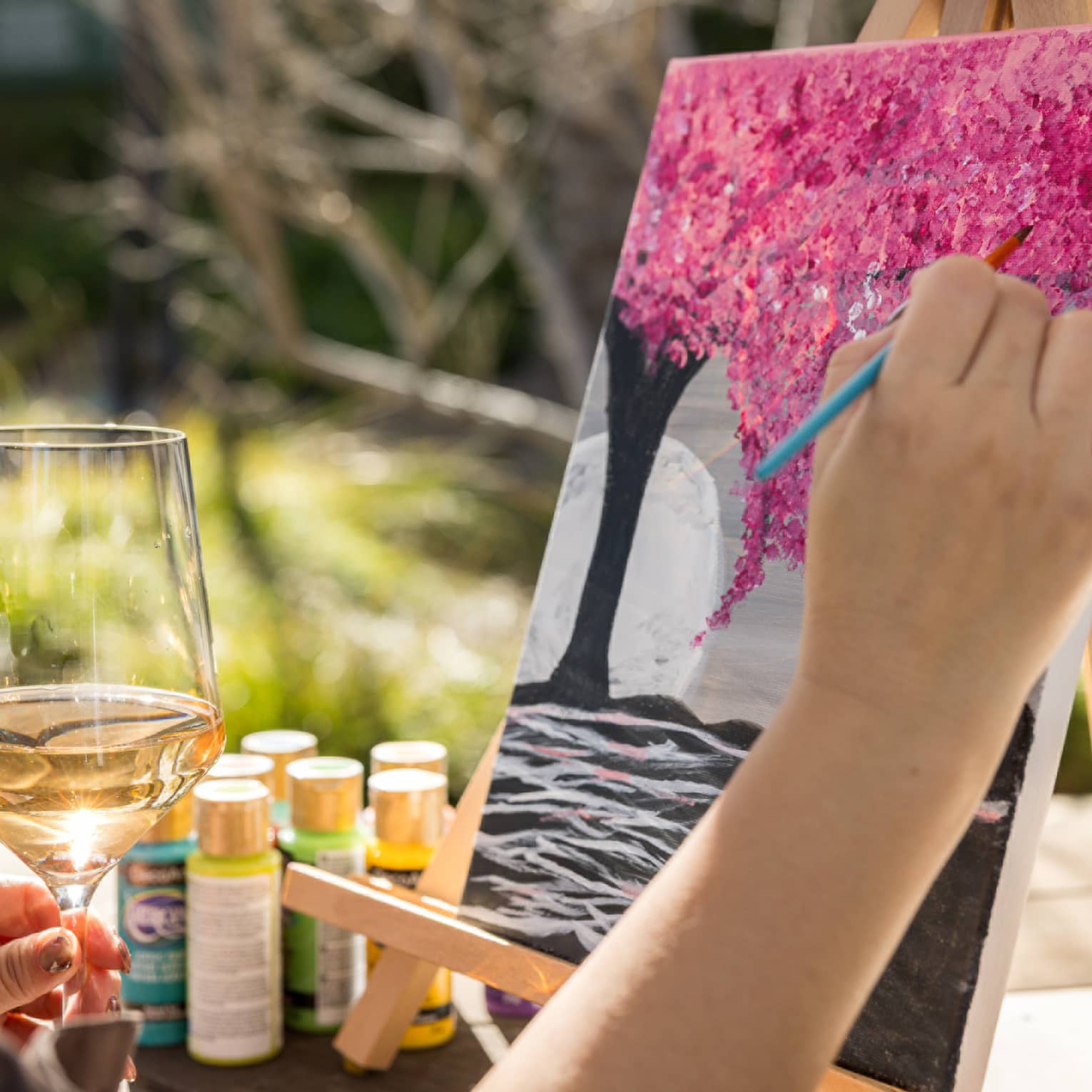 Person painting a pink tree on a canvas with one hand while holding a glass of wine in their other hand