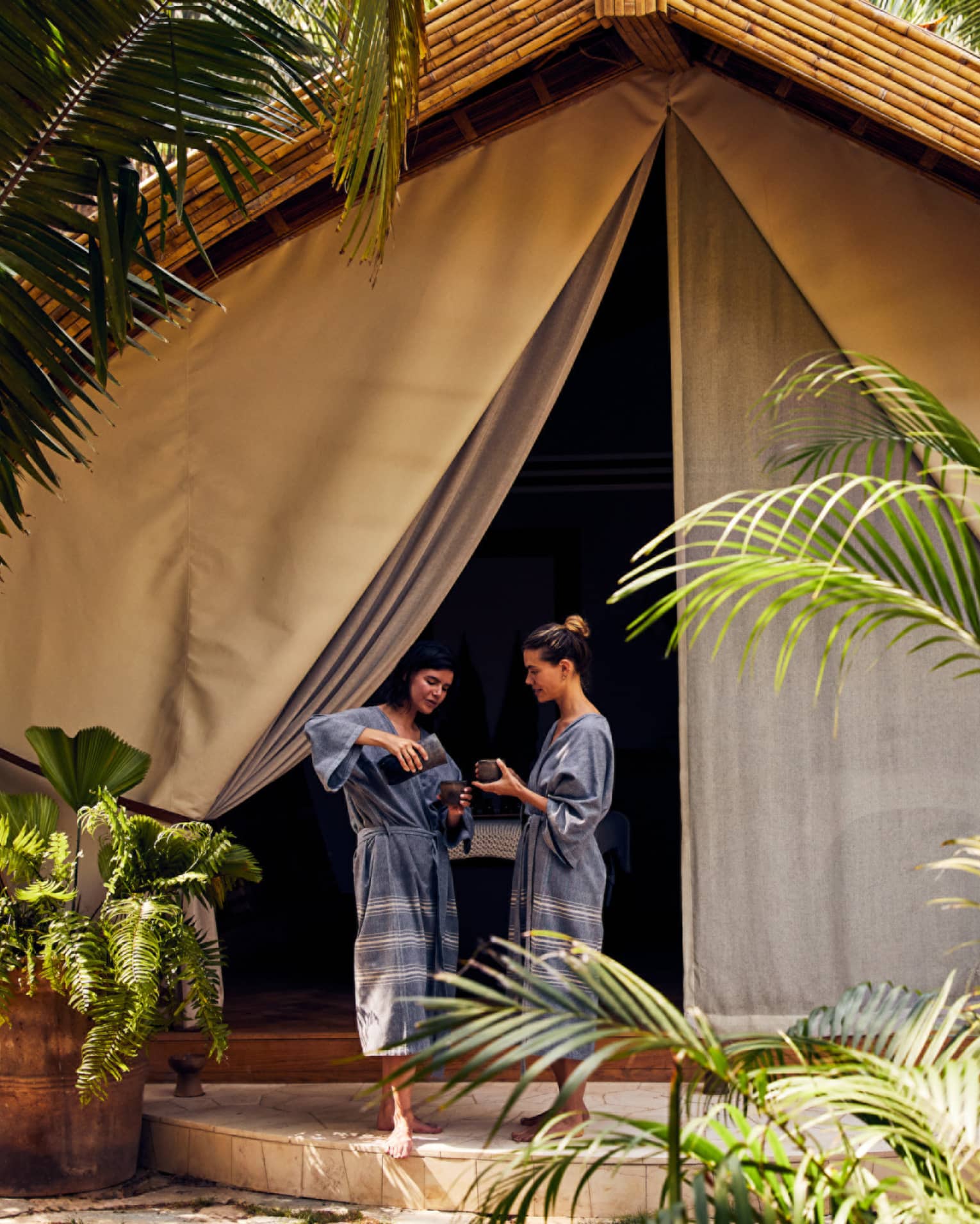 Two women in spa robes stand outside a luxury spa tent, one pouring tea for the other, surrounded by lush greenery in a peaceful setting.
