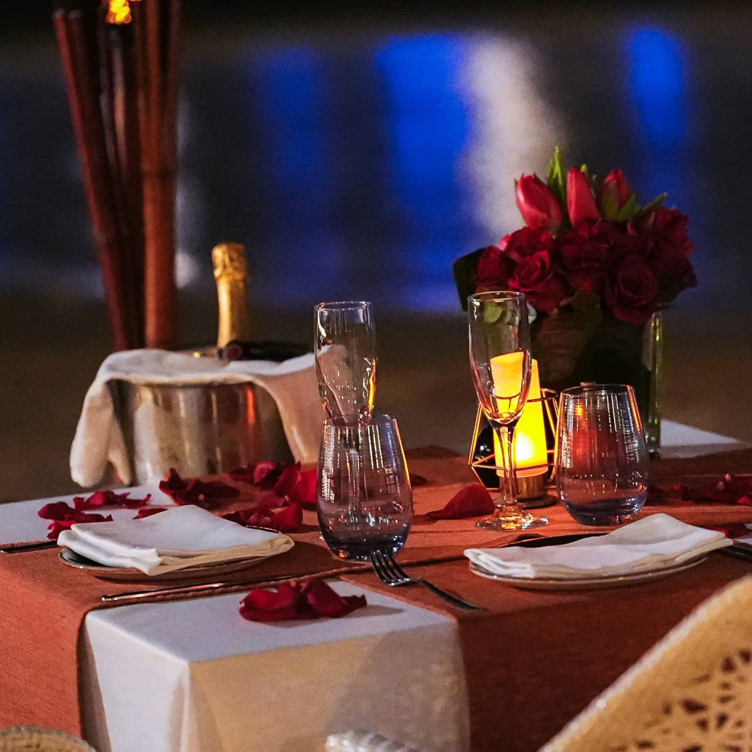 Table for two set with chilling champagne, flutes and a rose bouquet, aside torches and the gleaming reflection on the water.