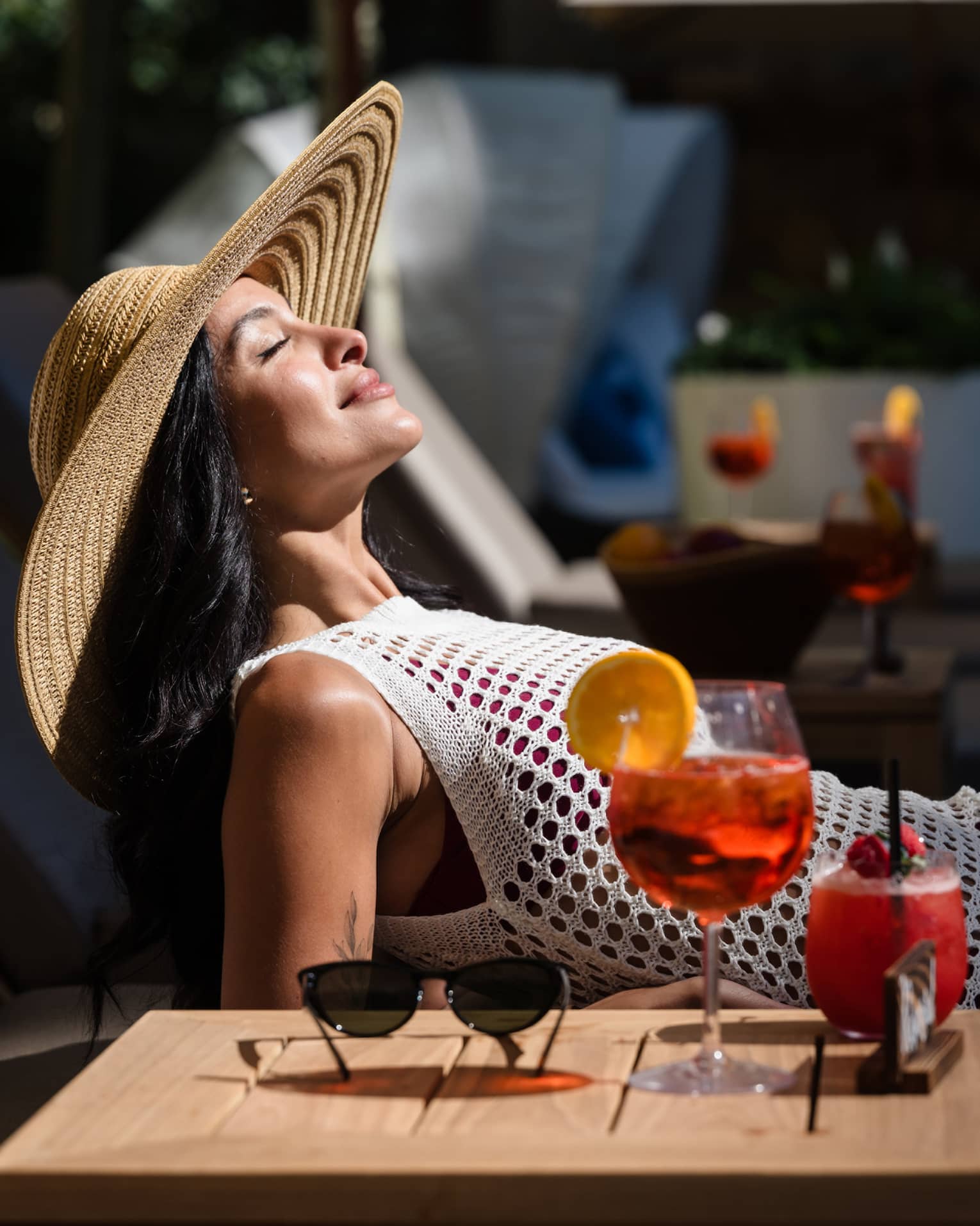 A guest laying in a lounge chair in the sunshine with their sunglasses and drink on a table beside them