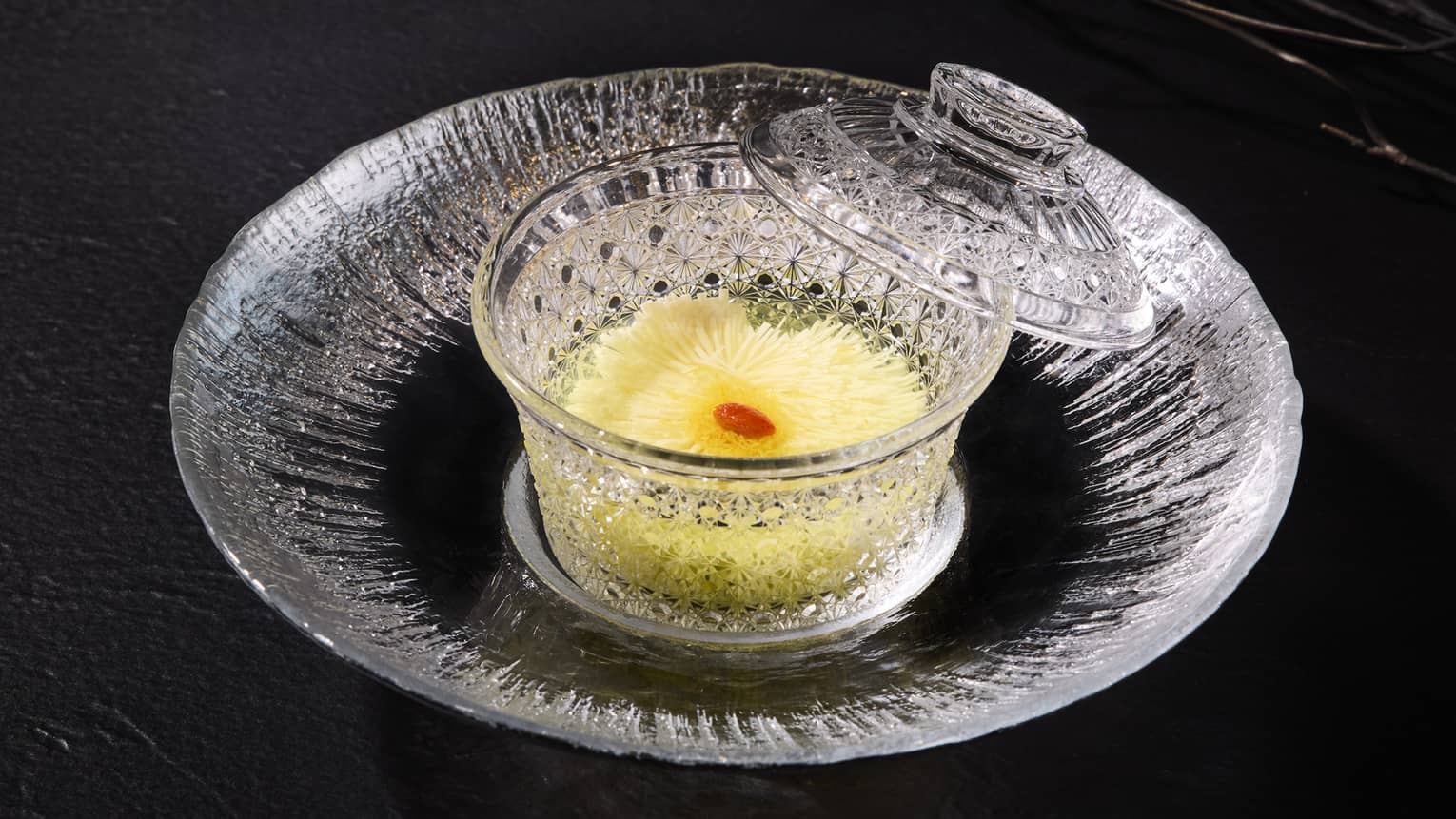 A clear glass plate with a clear glass bowl in the center holds a yellow food item with a small red garnish on top; the bowl?s clear glass lid is propped on the plate against the bowl?s edge.
