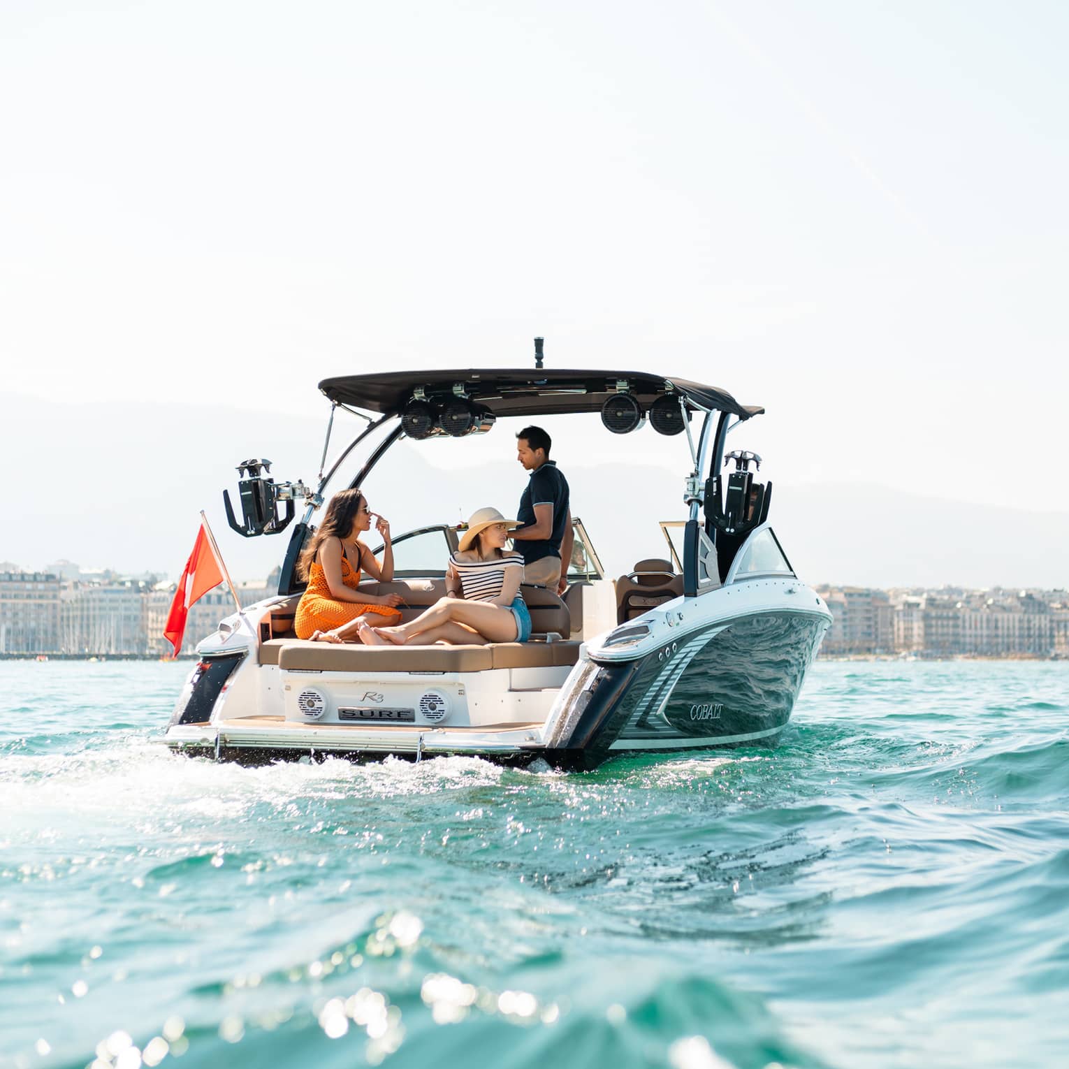 Captain guides a boat toward a building-lined shore while two guests lounge on a large cushioned seat behind him.