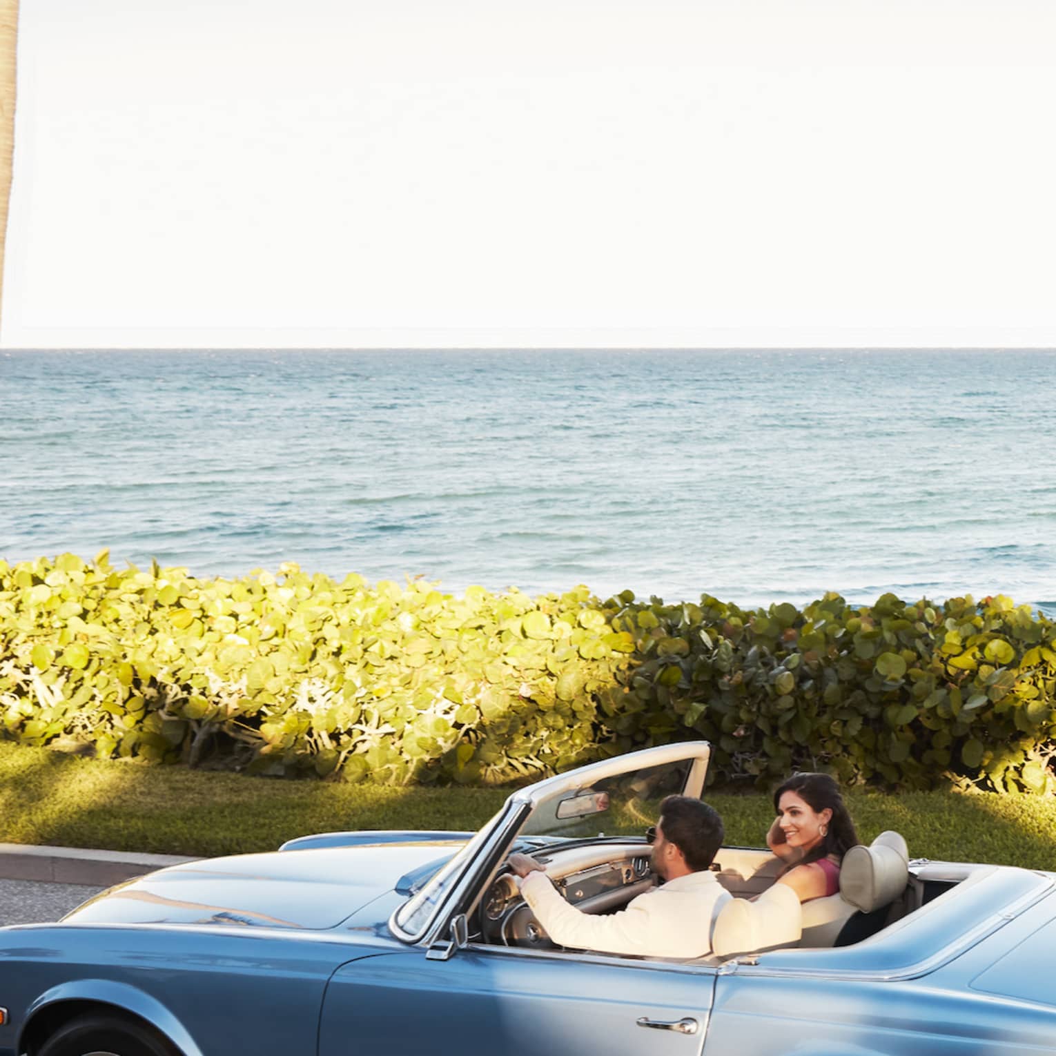 Two people in a convertible car driving down a road next to the ocean.