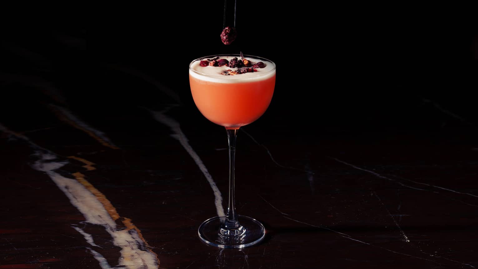 A hand reaches out of the darkness to drop dried cranberries onto a red-colored cocktail in a tall stemmed glass