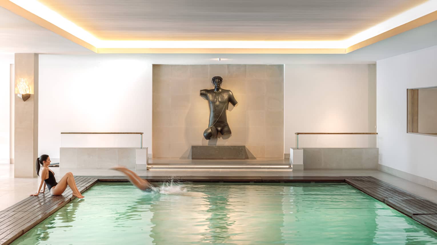 An indoor pool in a bright space finished in neutral tones. A figurative sculpture is displayed against a wall at the end of the pool.