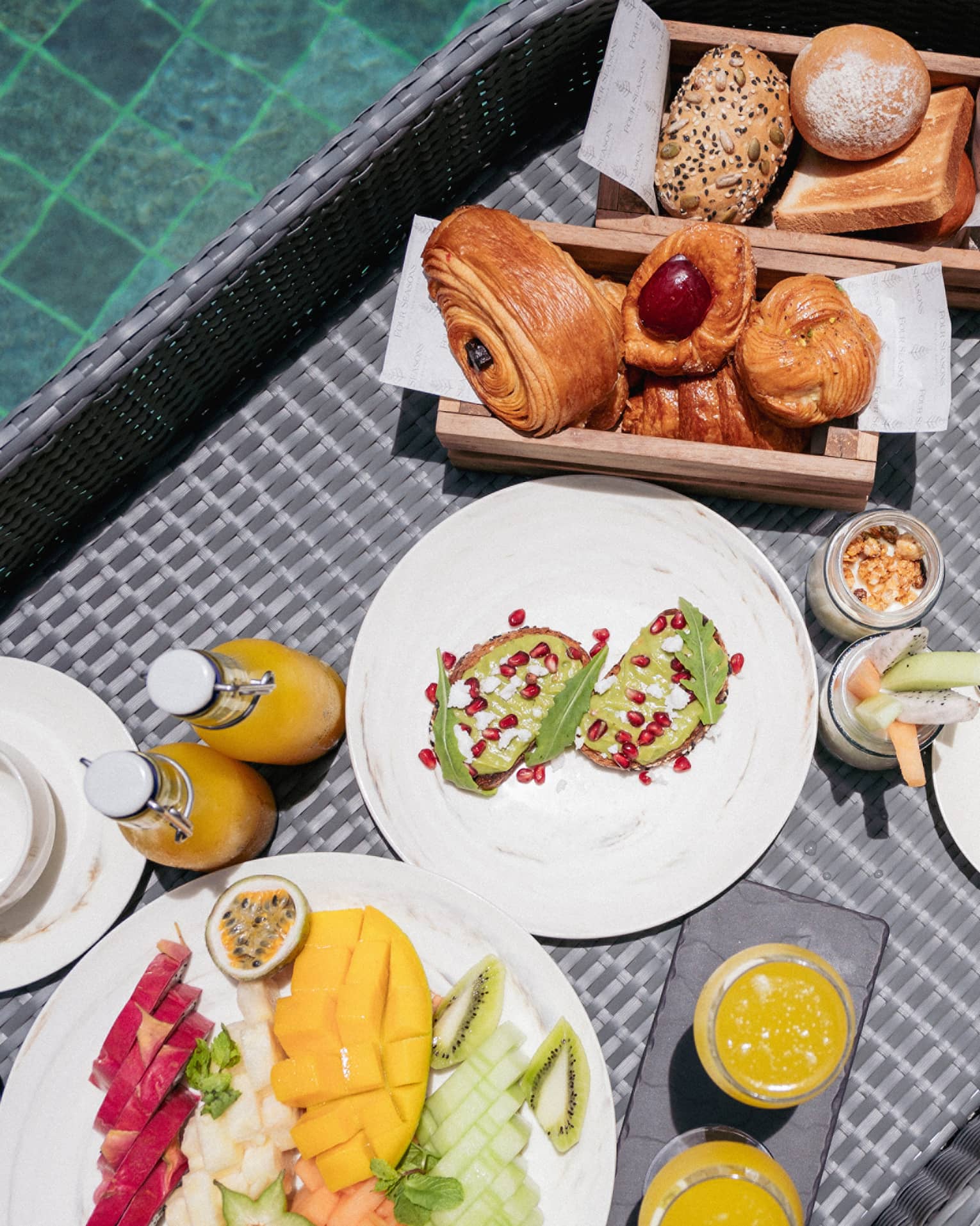 Close-up view from above of a wicker tray filled with fruit, pastries, juice and toast floating in a crystal-clear pool.