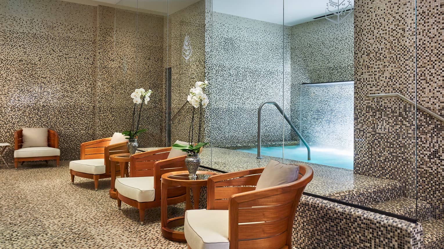 Tile spa room with whirlpool bath behind row of wood chairs with white cushions, tables with white orchids