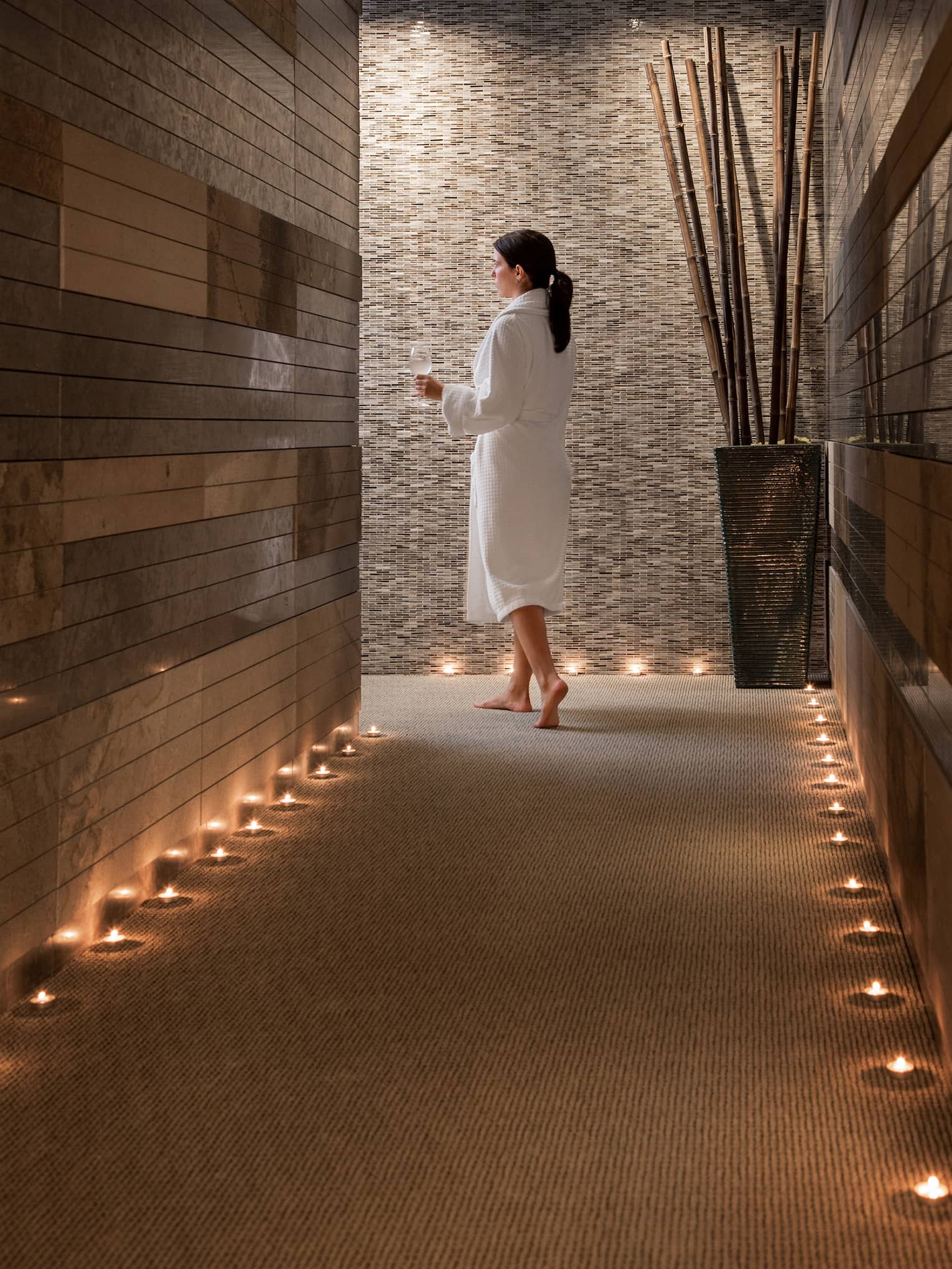 Woman in bathrobe holding wine glass with water walks through spa