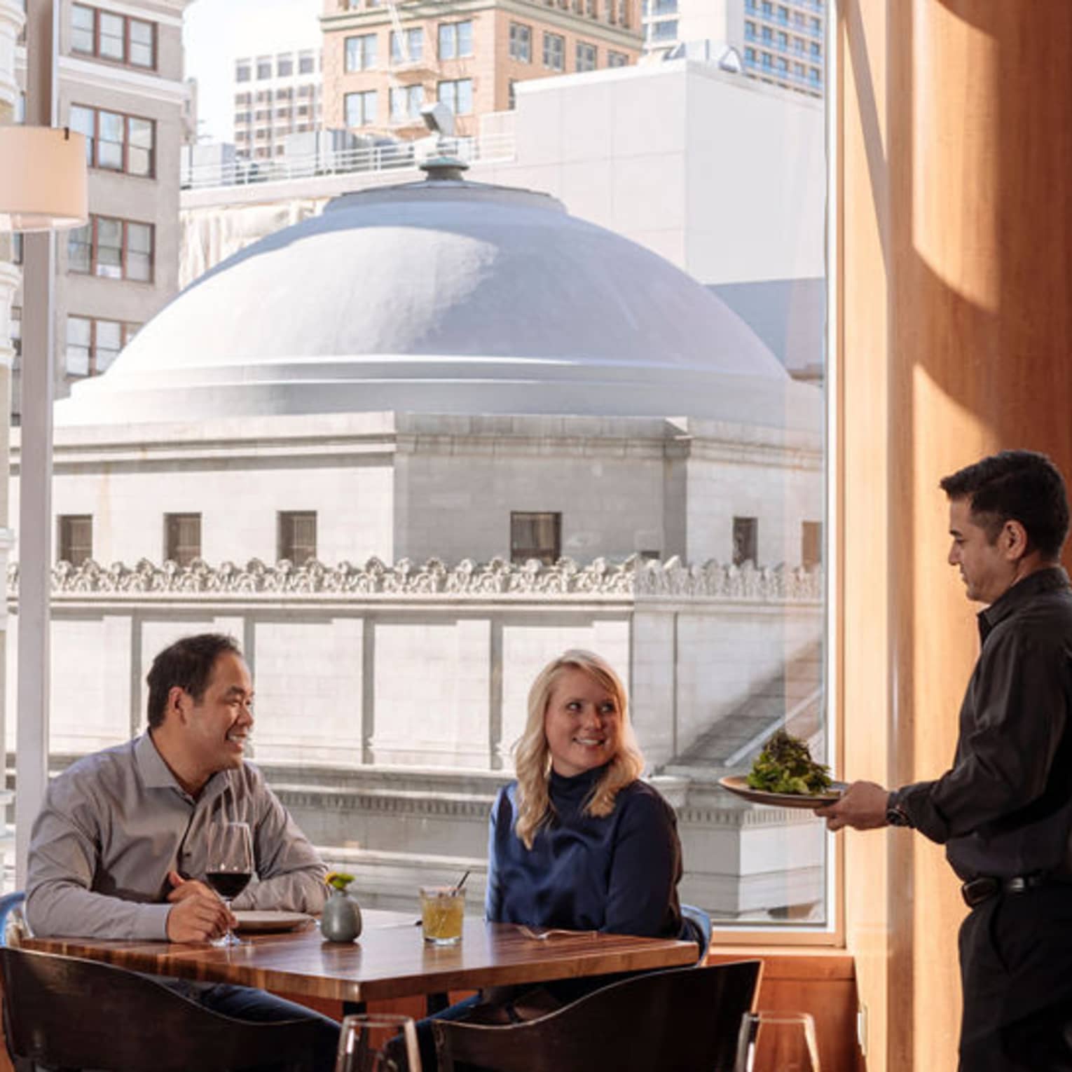 A four seasons staff takes a couple's order as they sit at a window table overlooking the city 