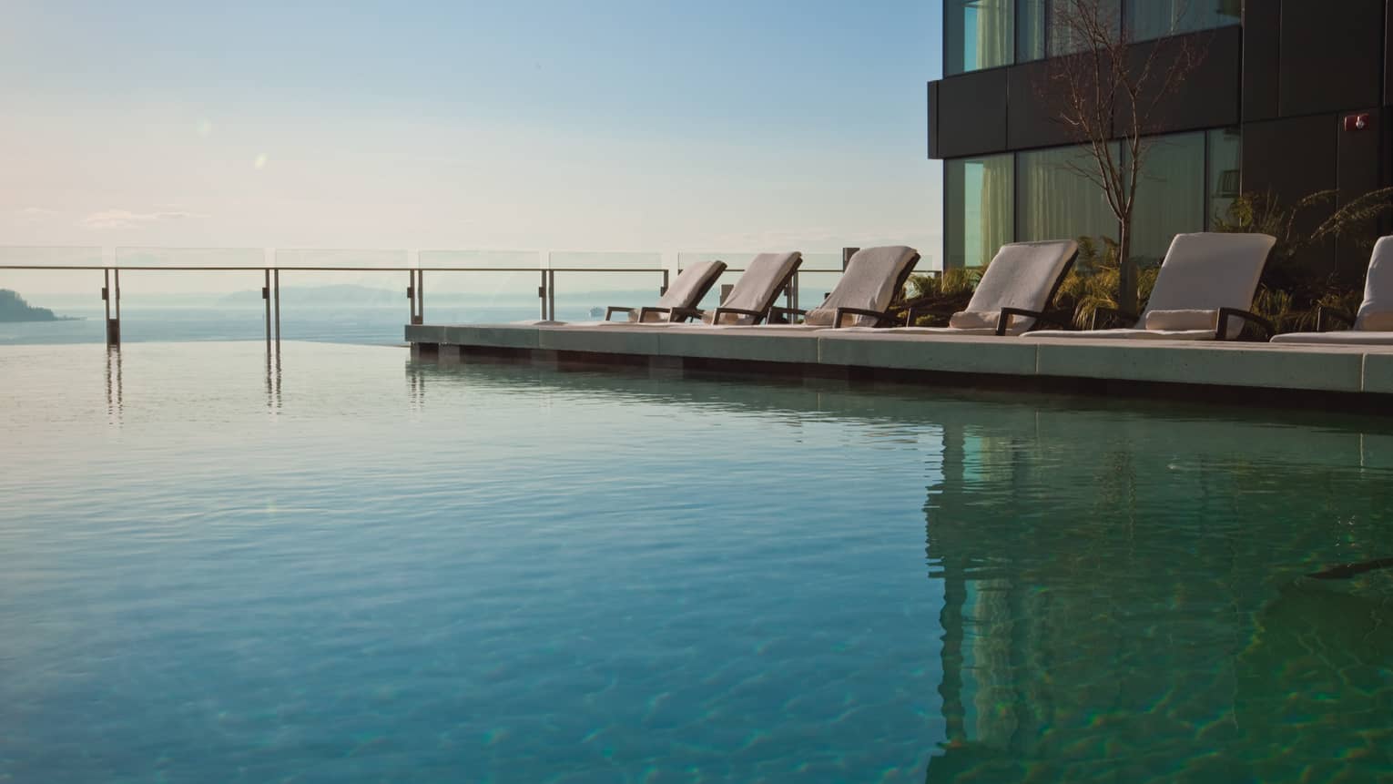 Deck chairs on patio over turquoise waters of outdoor infinity swimming pool by ocean