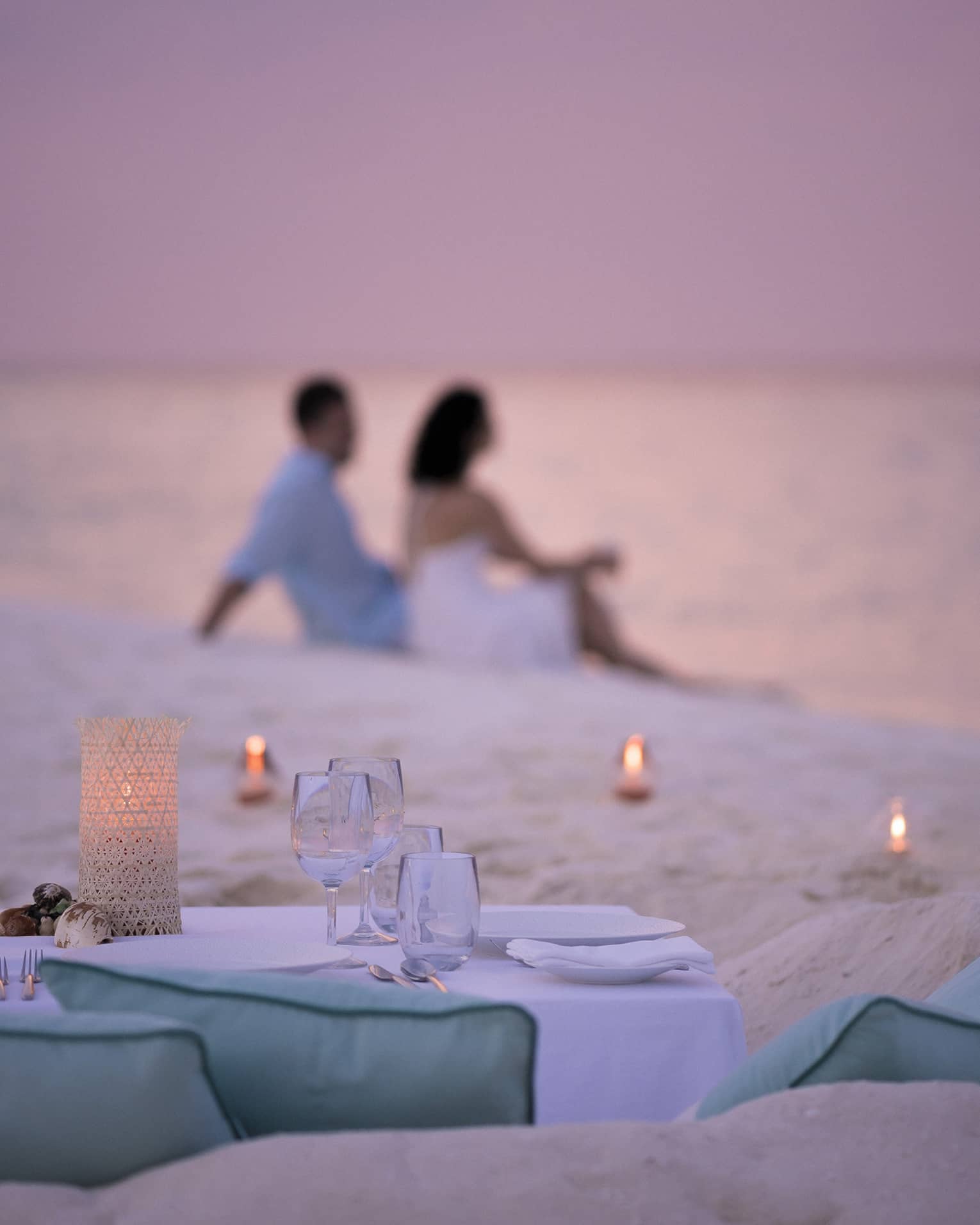 Private dining table in front of sand beach with tealight candles, couple sitting in sand