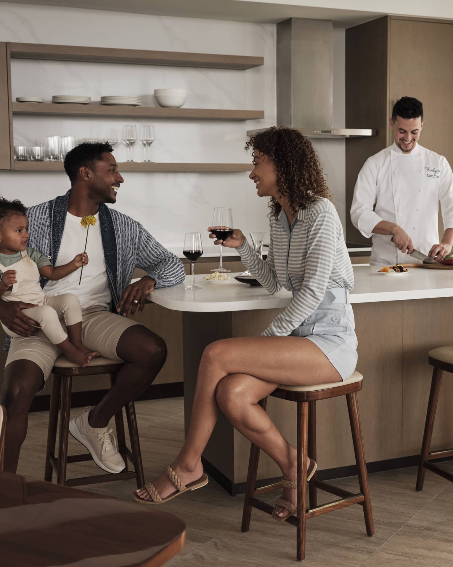 Parents sit at a kitchen island, toddler in dad's arms, laughing over glasses of red wine while a chef prepares a meal. 