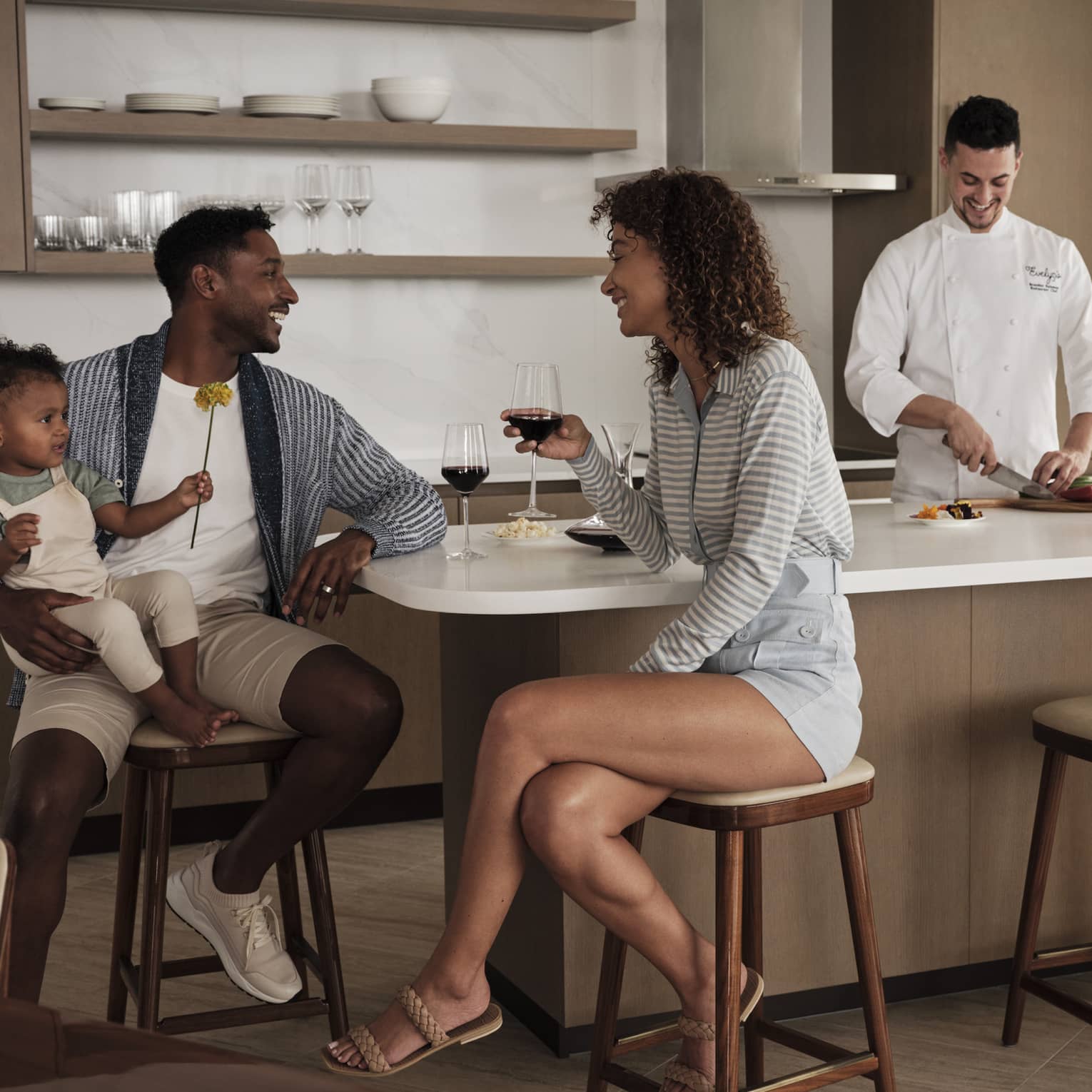 Parents sit at a kitchen island, toddler in dad's arms, laughing over glasses of red wine while a chef prepares a meal. 