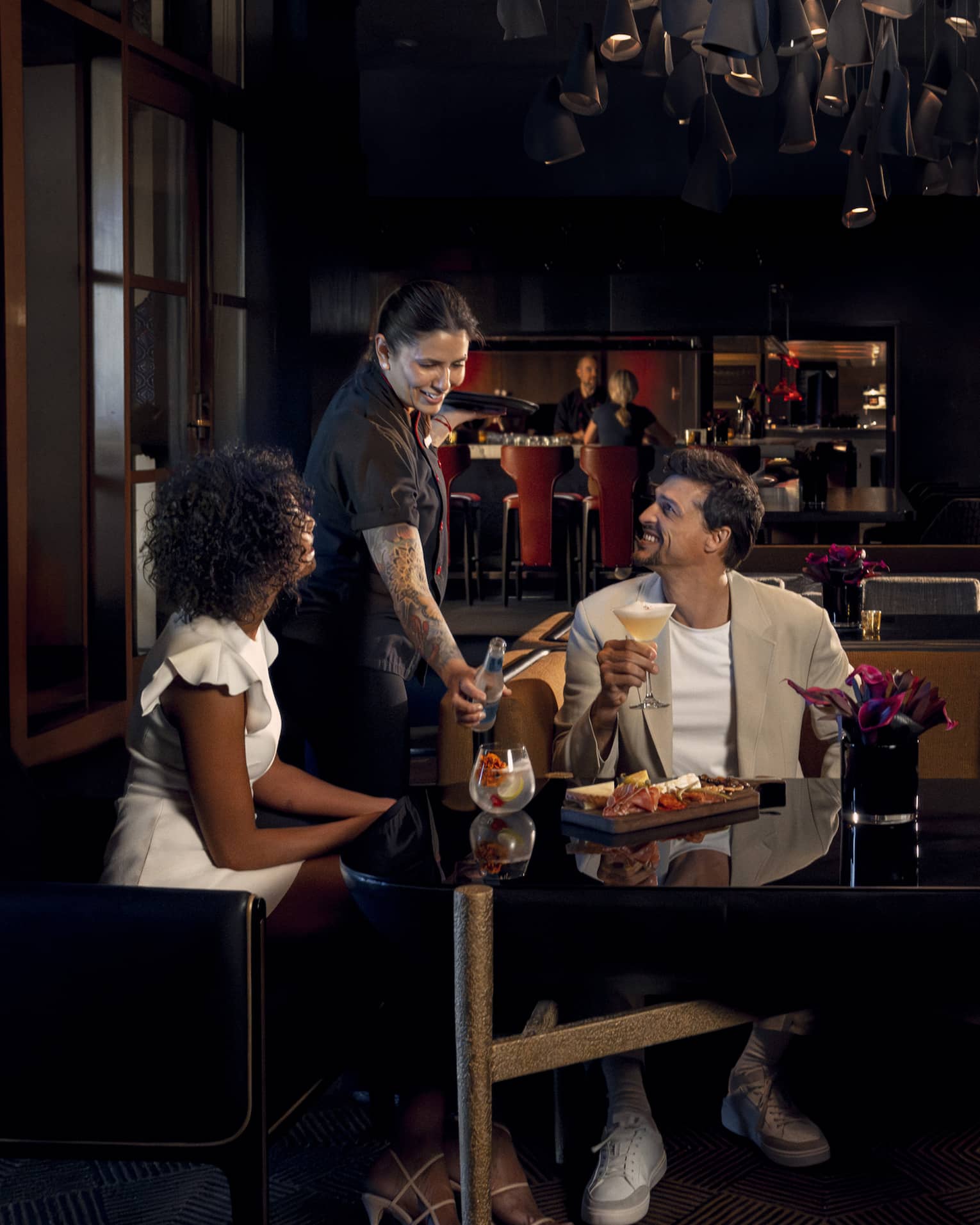 Two people wearing light-coloured cocktail attire sit on benches around a black table as a waitress brings them beverages