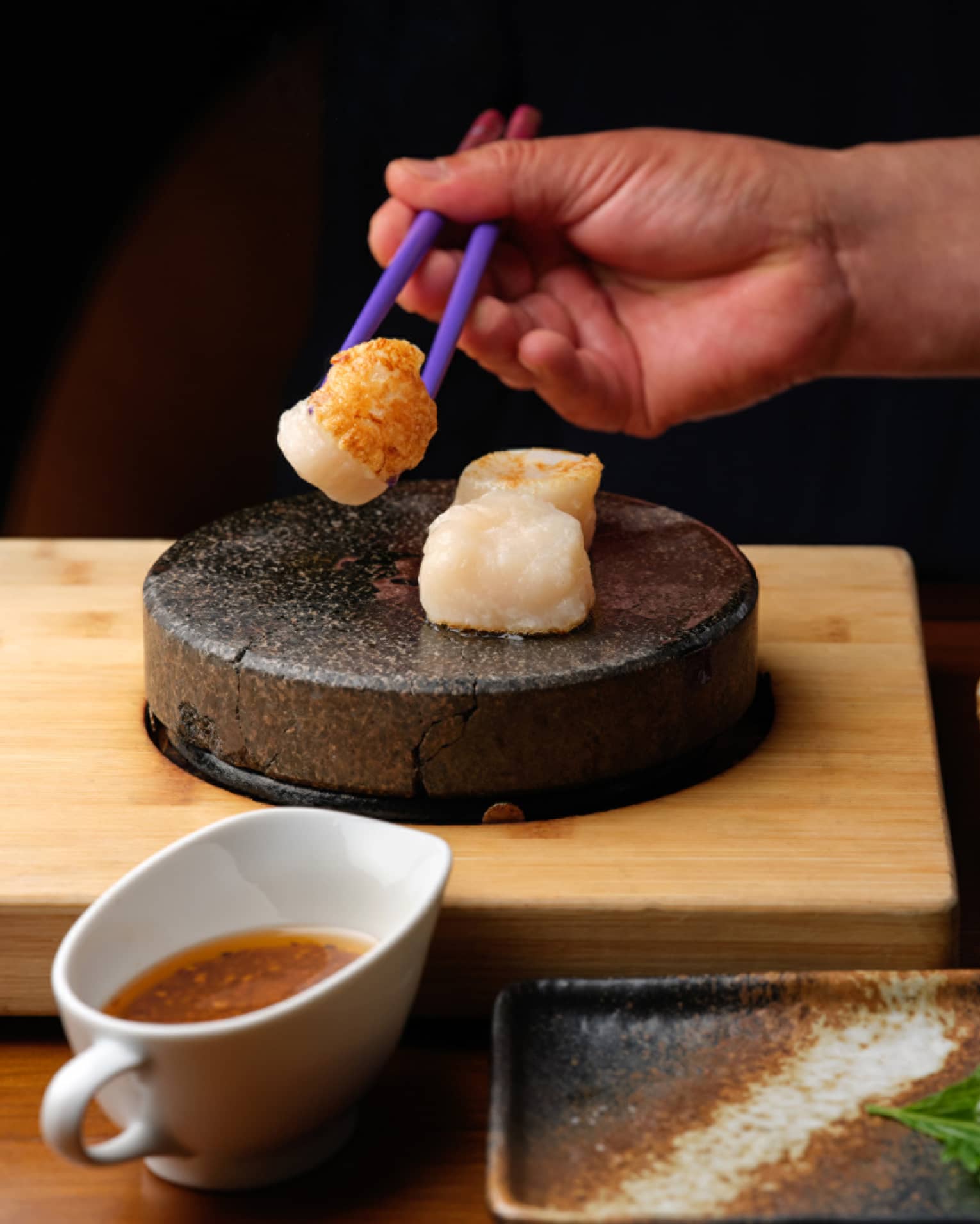 Close-up of a hand picking up a seared scallop with chopsticks from a stone tray, near small dishes of sauce and fresh sides.
