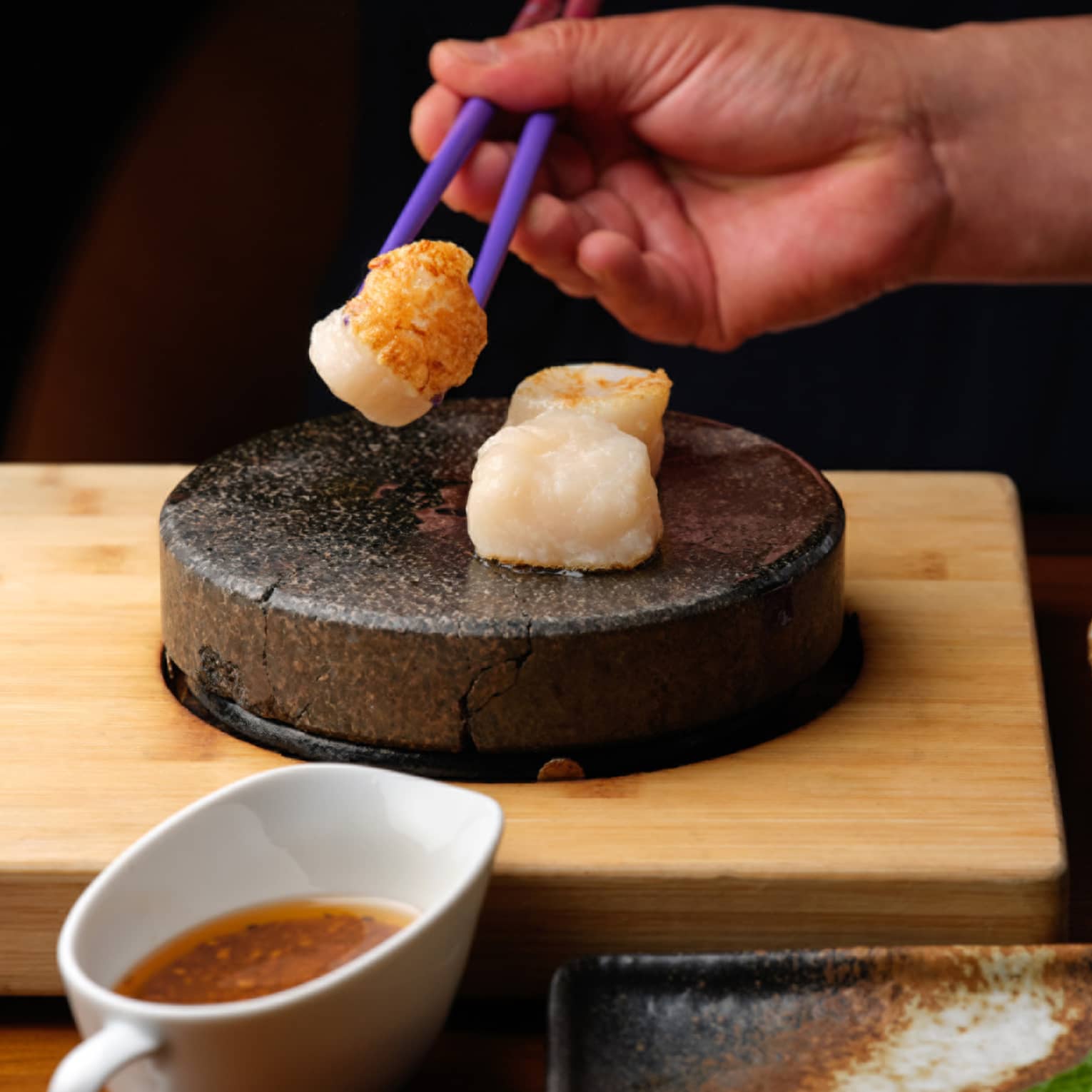 Close-up of a hand picking up a seared scallop with chopsticks from a stone tray, near small dishes of sauce and fresh sides.