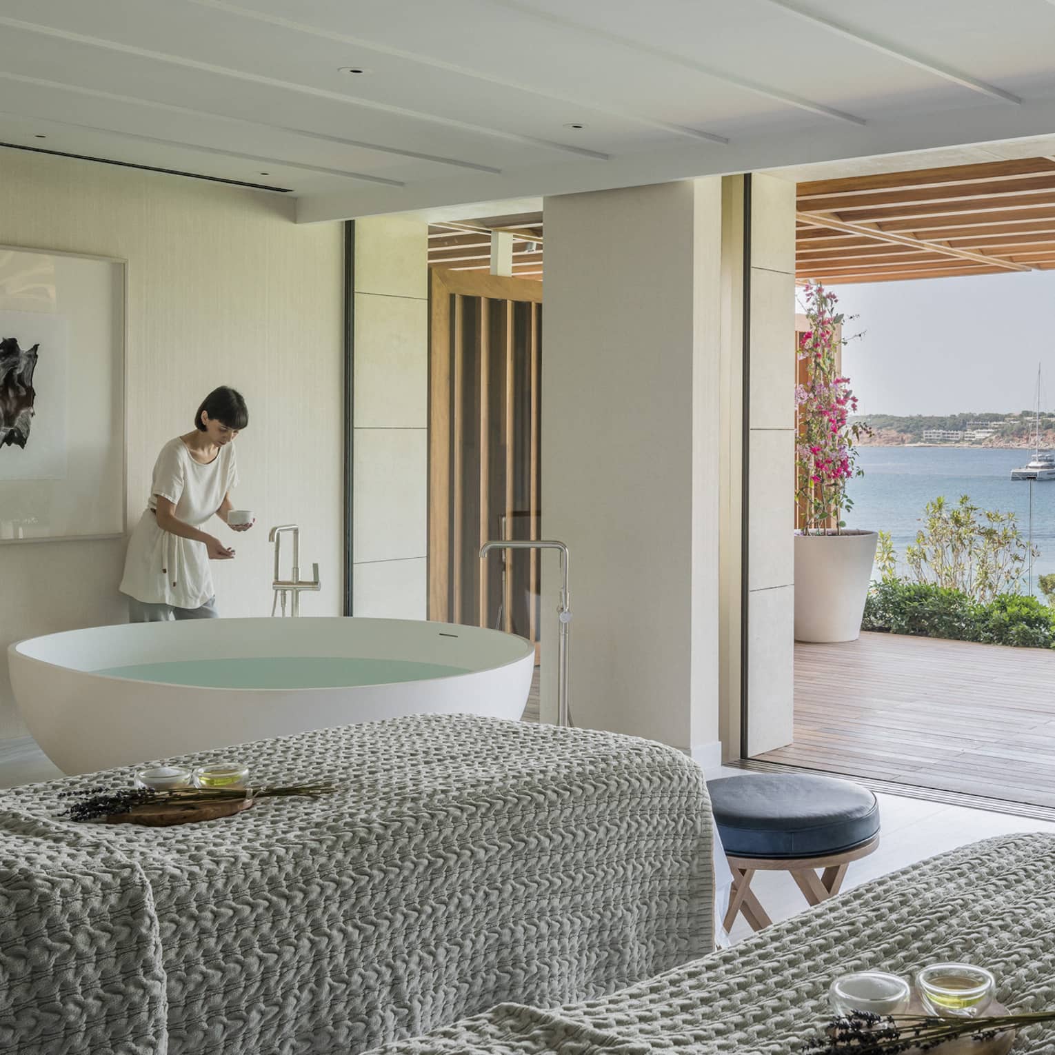 A four seasons staff works in a room with two side by side spa tables draped with gray linens face an open veranda overlooking a body of water in Greece