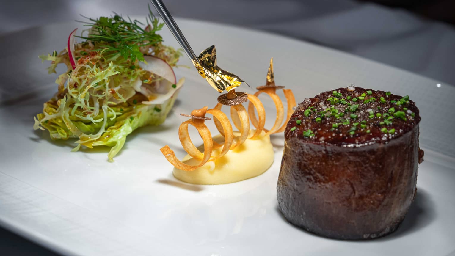 Beef tenderloin, small bunch of salad and a curl-shapped side dish all plated in a neat line on a white rectangular plate