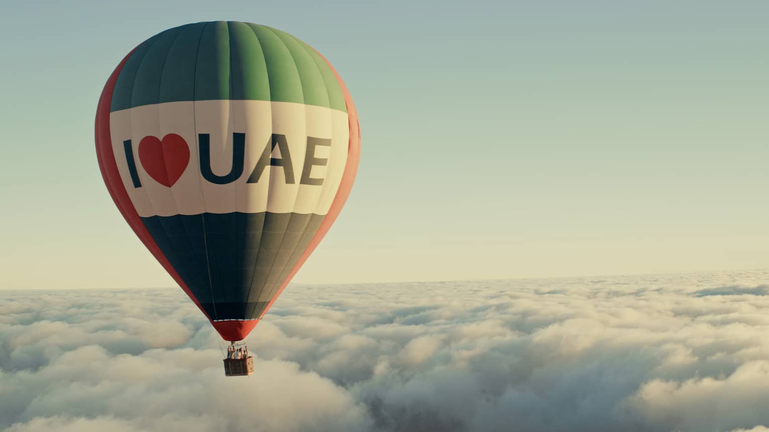 A hot air balloon flies above the clouds, in front of a light blue sky.