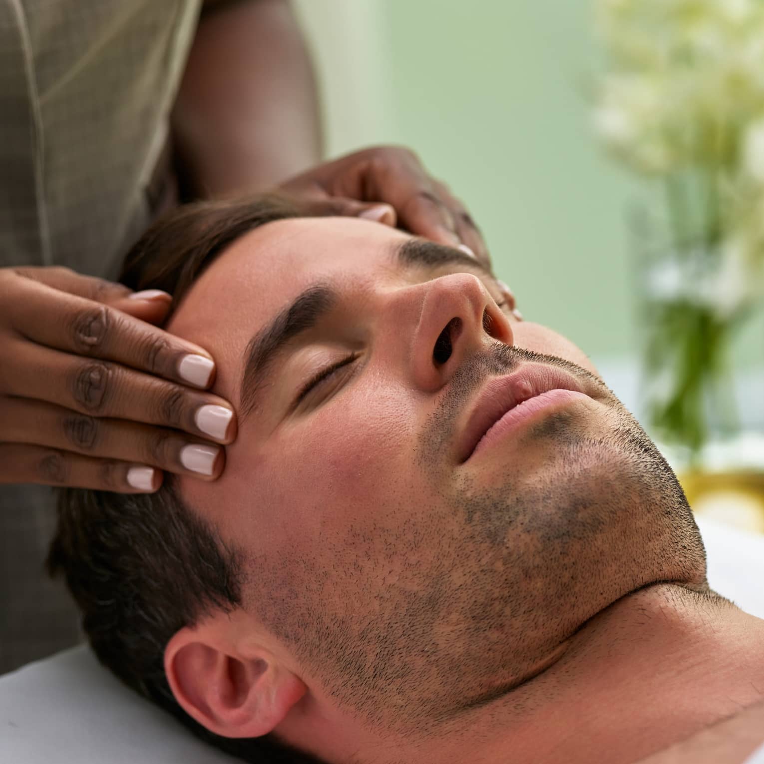 Man lies on massage table with eyes closed as hands massage his temples, forehead