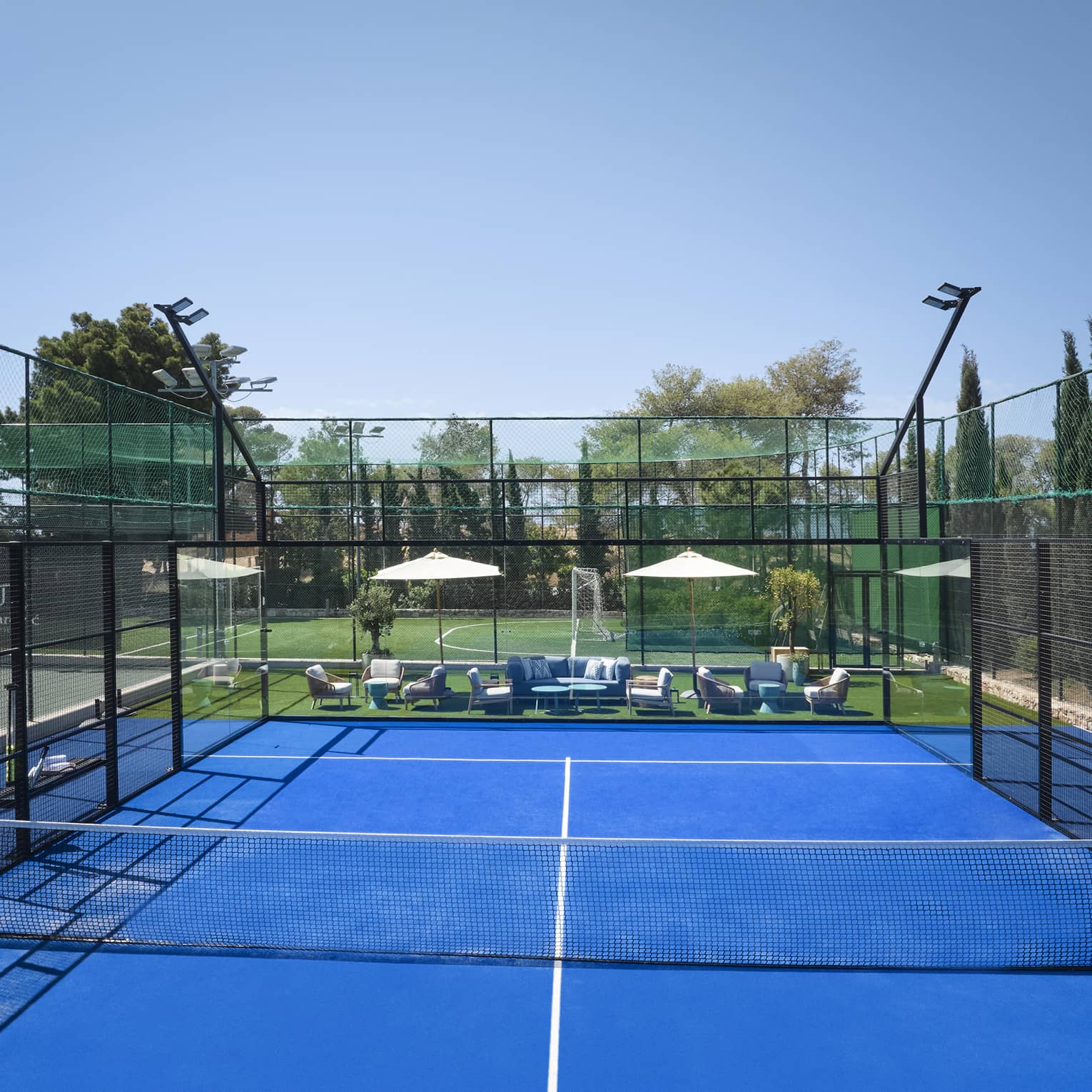 Bright blue padel court surrounded by fencing, white umbrellas and lounge chairs