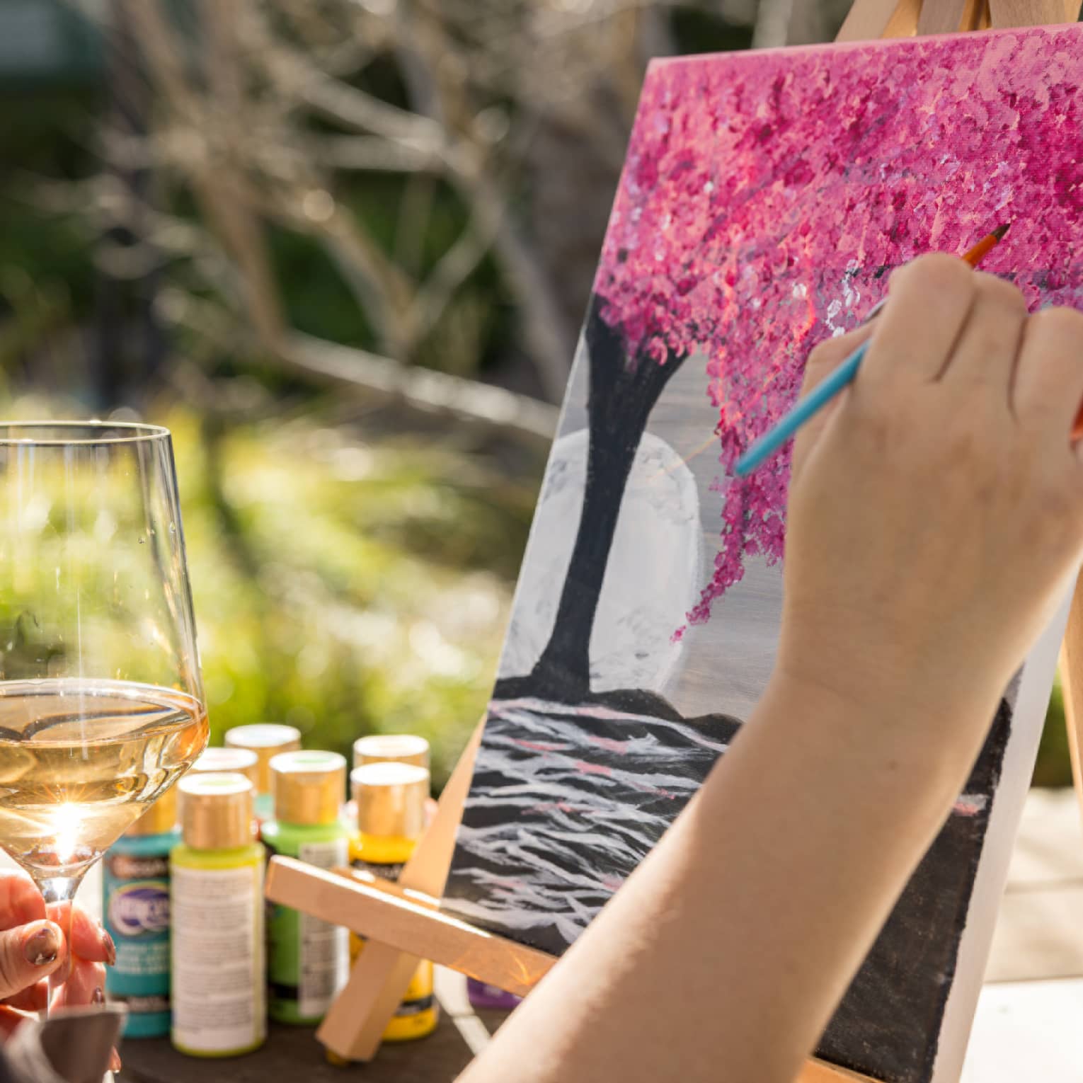 Person painting a pink tree on a canvas with one hand while holding a glass of wine in their other hand