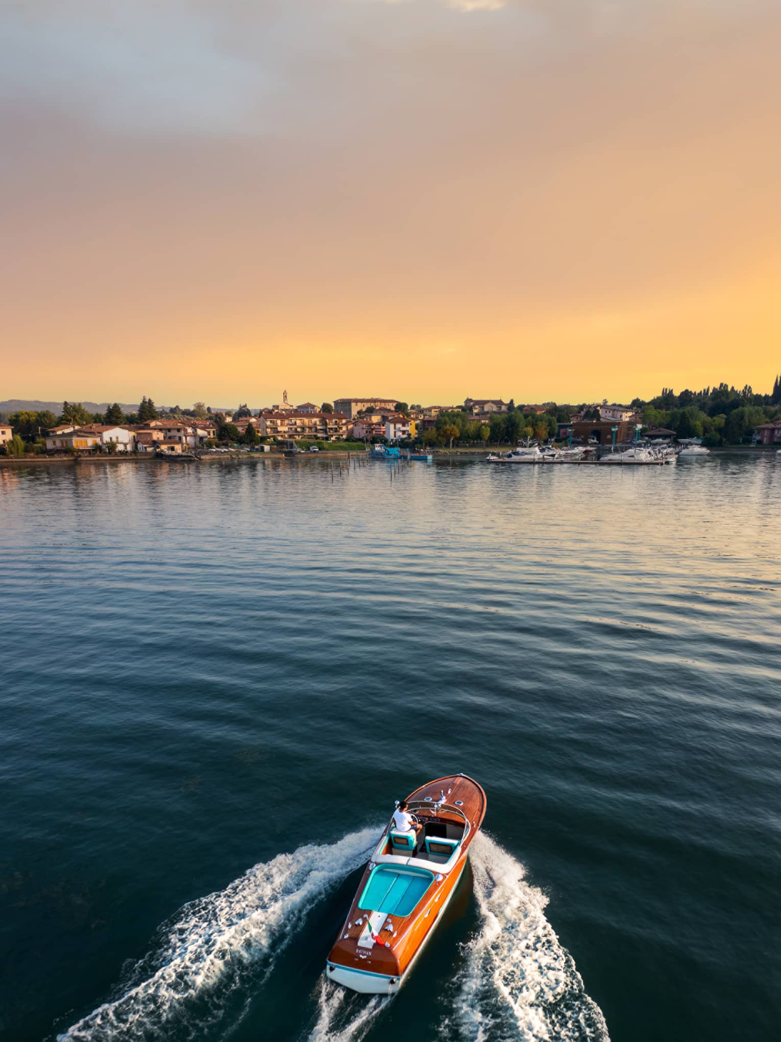 Person driving a wooden speedboat across a lake towards shore at sunset