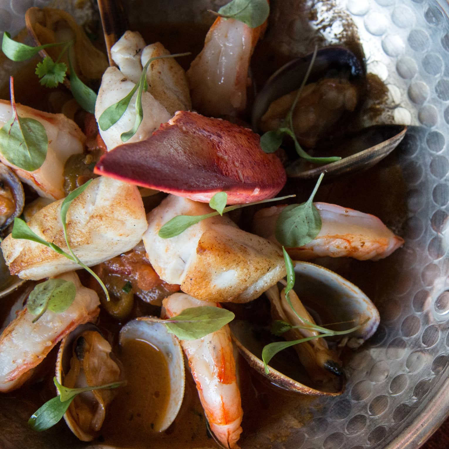 Close-up of a patterned metal bowl filled with prawns, scallops and mussels in a rich, dark broth garnished with green herbs.