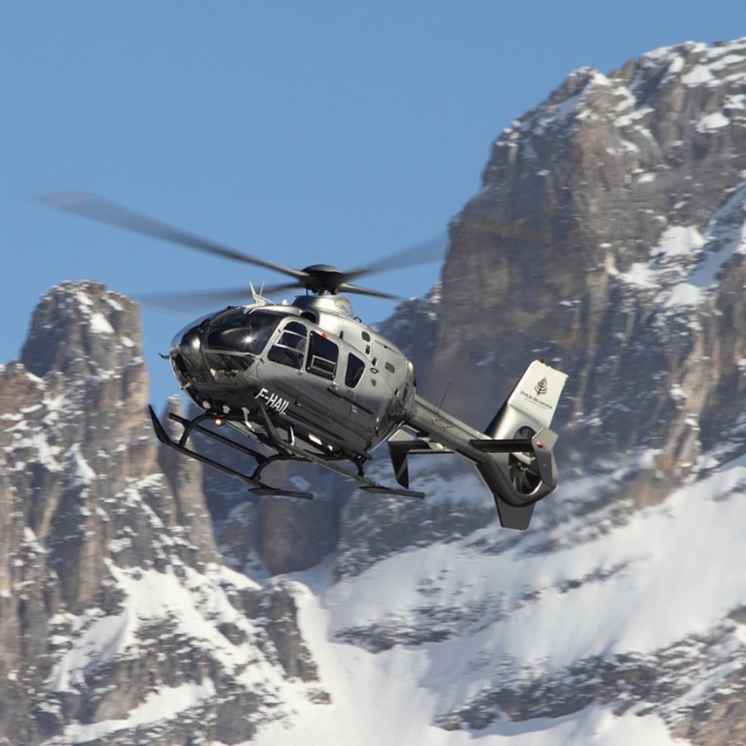 Silver helicopter ascending beside snowy, rocky mountain peaks