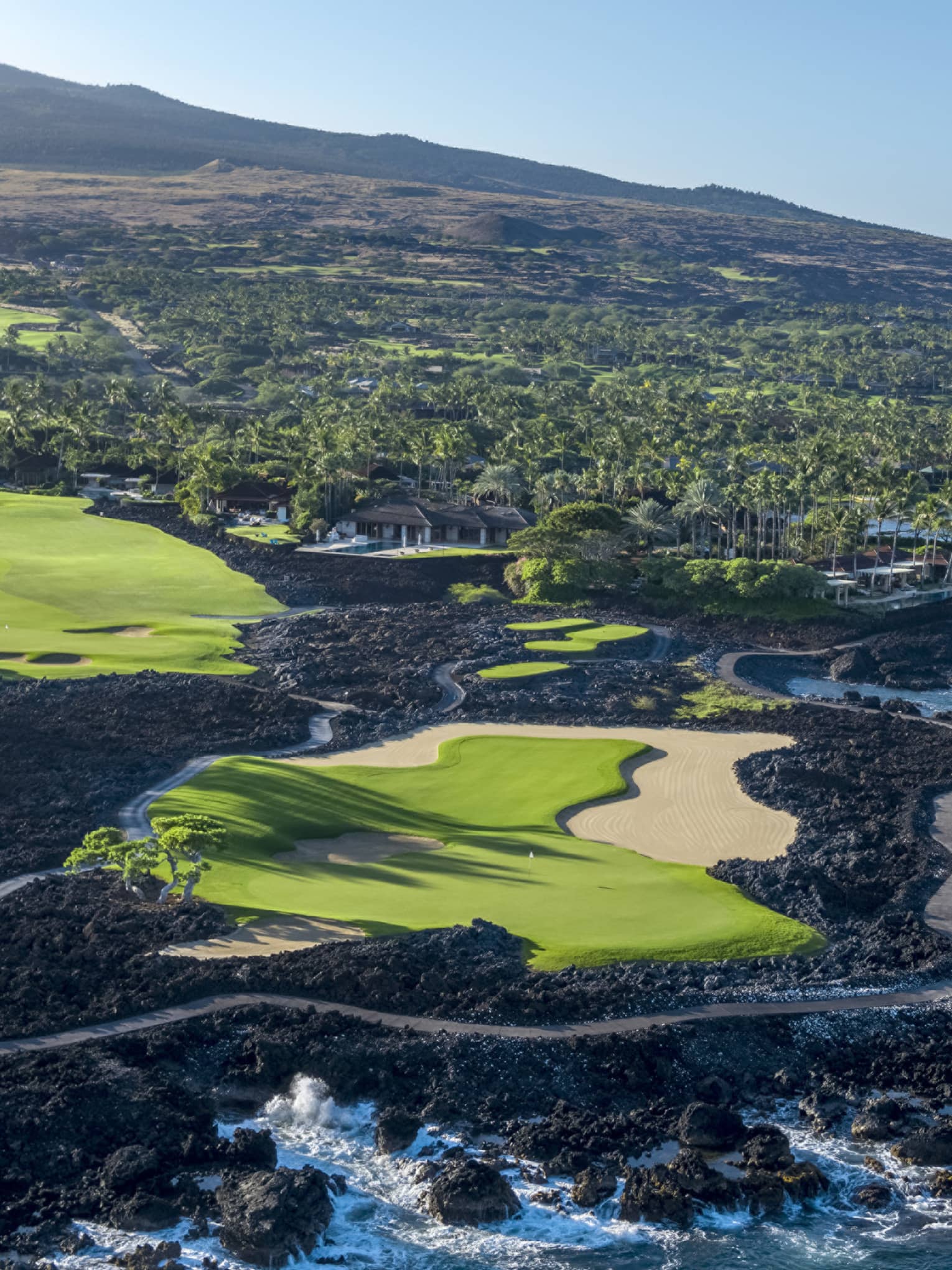 A lush green golf course lined with black lava, verdant forest, and azure ocean views