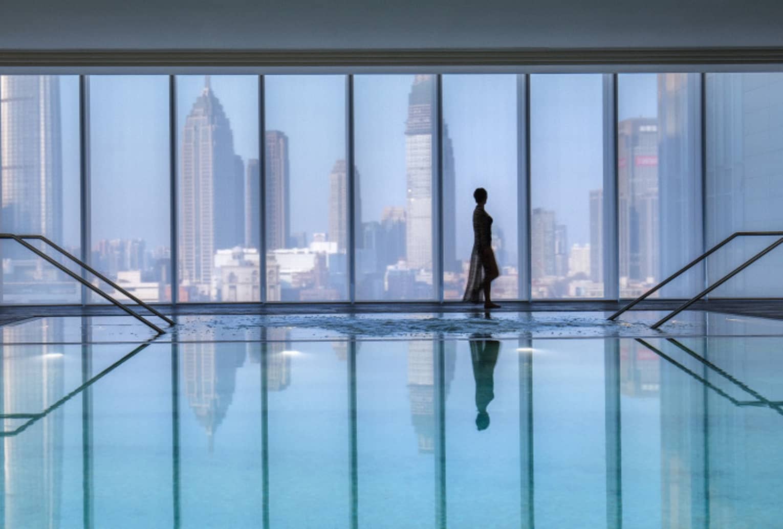 Silhouette of woman walking by blue indoor swimming pool, floor-to-ceiling window