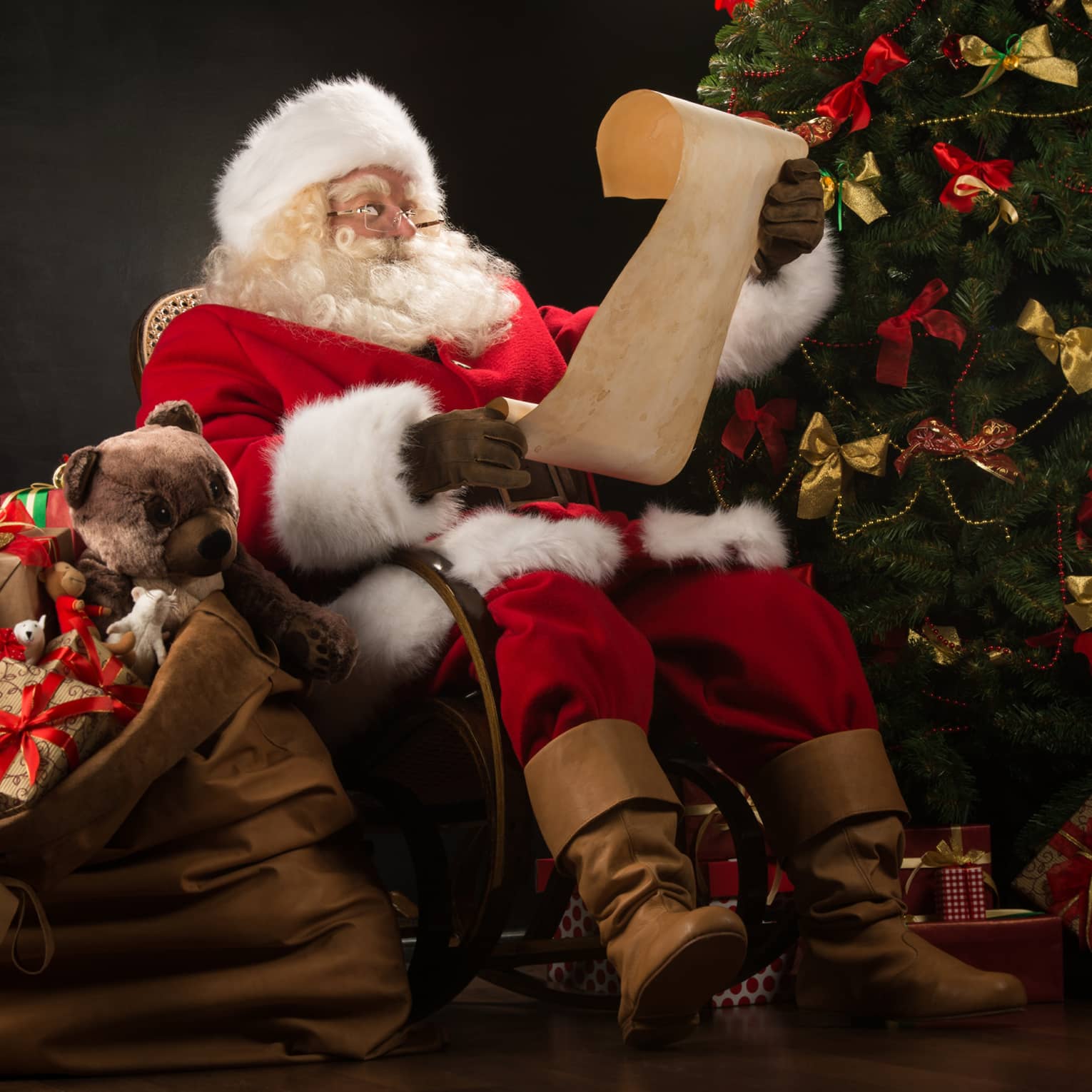 Santa Claus seated in large rocker holding a scroll, flanked by decorated Christmas tree with gifts and overflowing gift bag