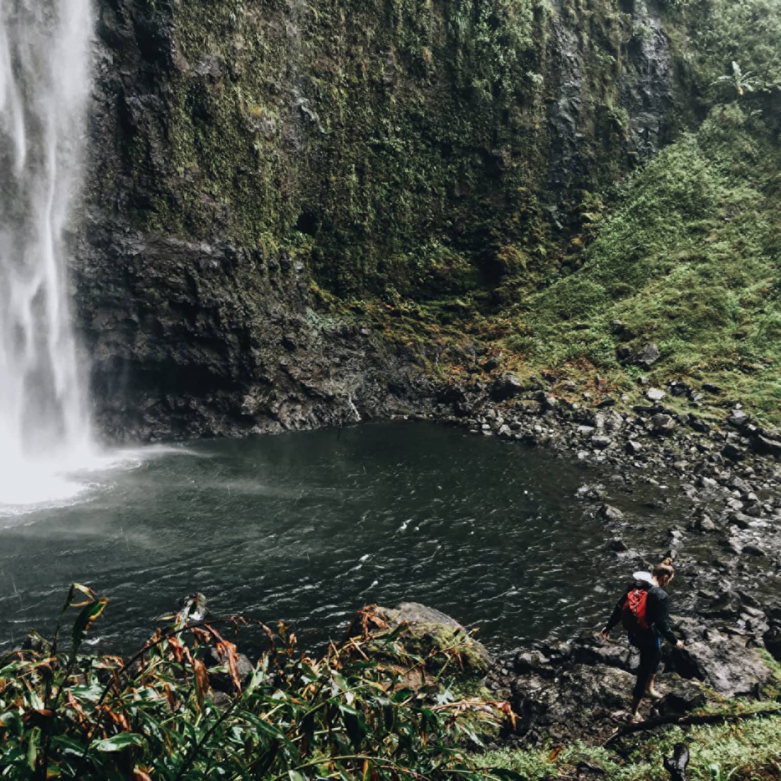 Back of woman hiking over black rocks under tall waterfall, small pond