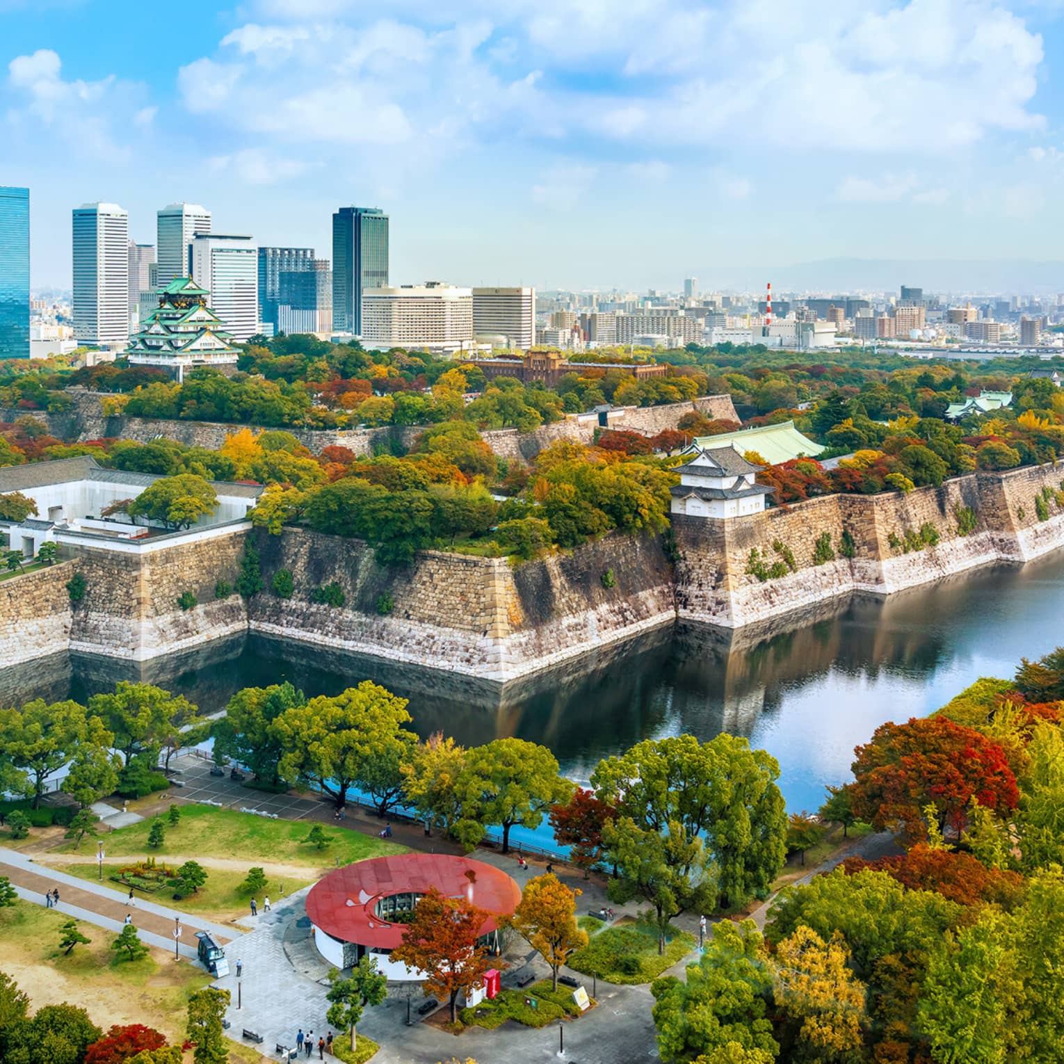 Osaka Castle rises amid lush autumn-coloured trees within a stone fortress surrounded by a moat and modern high-rises.
