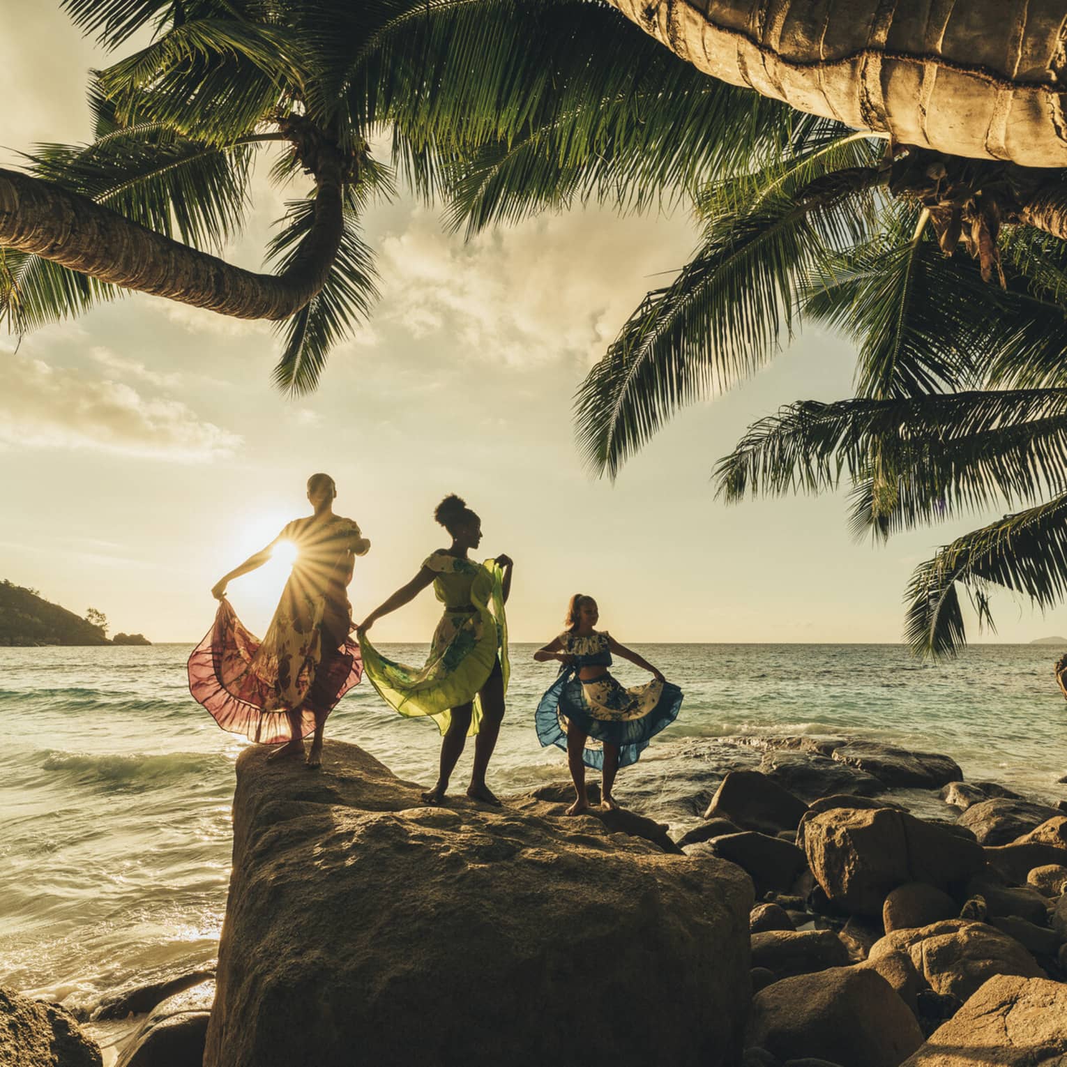 The sun sets over three dancers wearing long, colourful dresses standing on top of rocks by the water