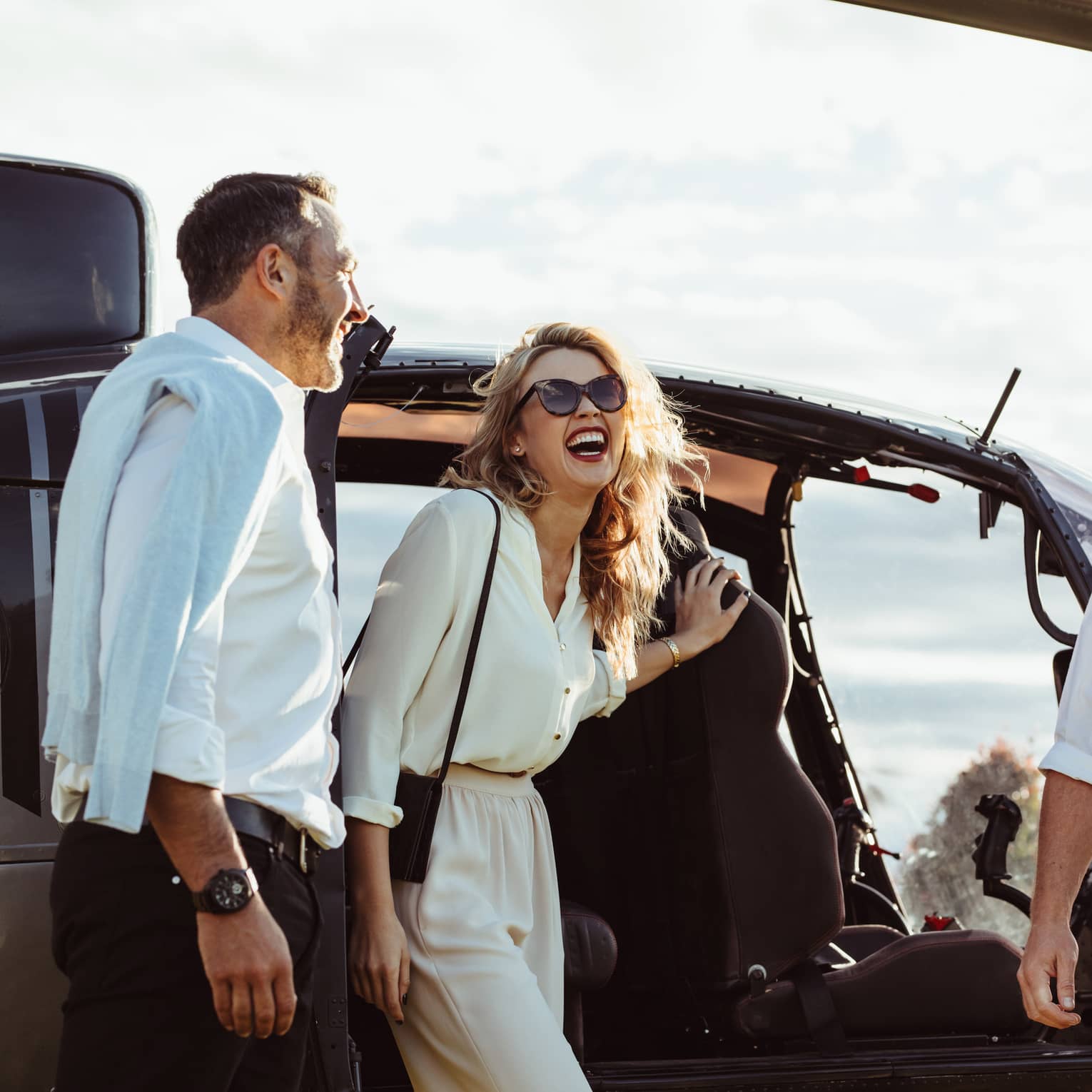 A man and woman laughing while getting out of a helicopter with the pilot smiling nearby.
