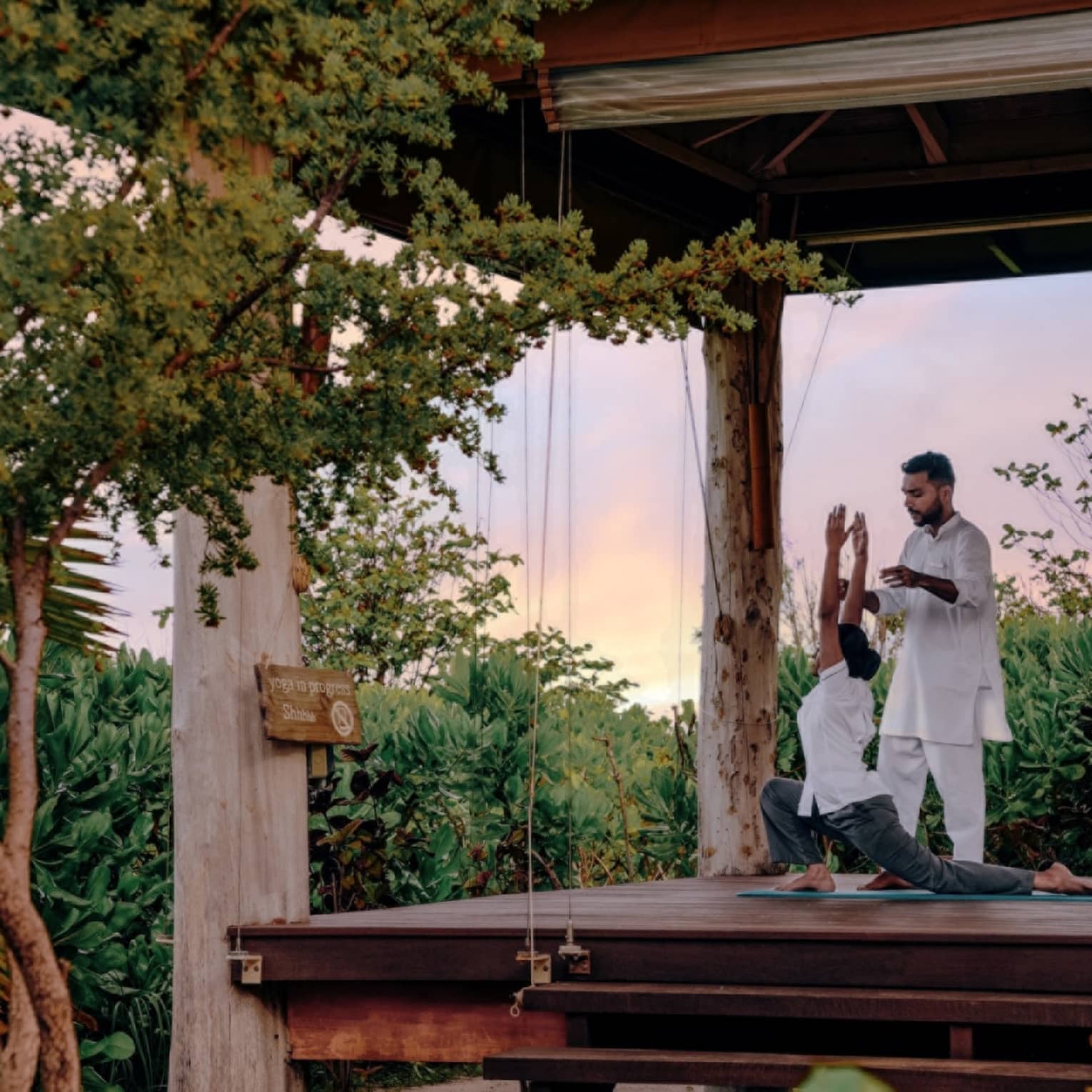Instructor guides student doing yoga in an open-air pavilion surrounded by greenery