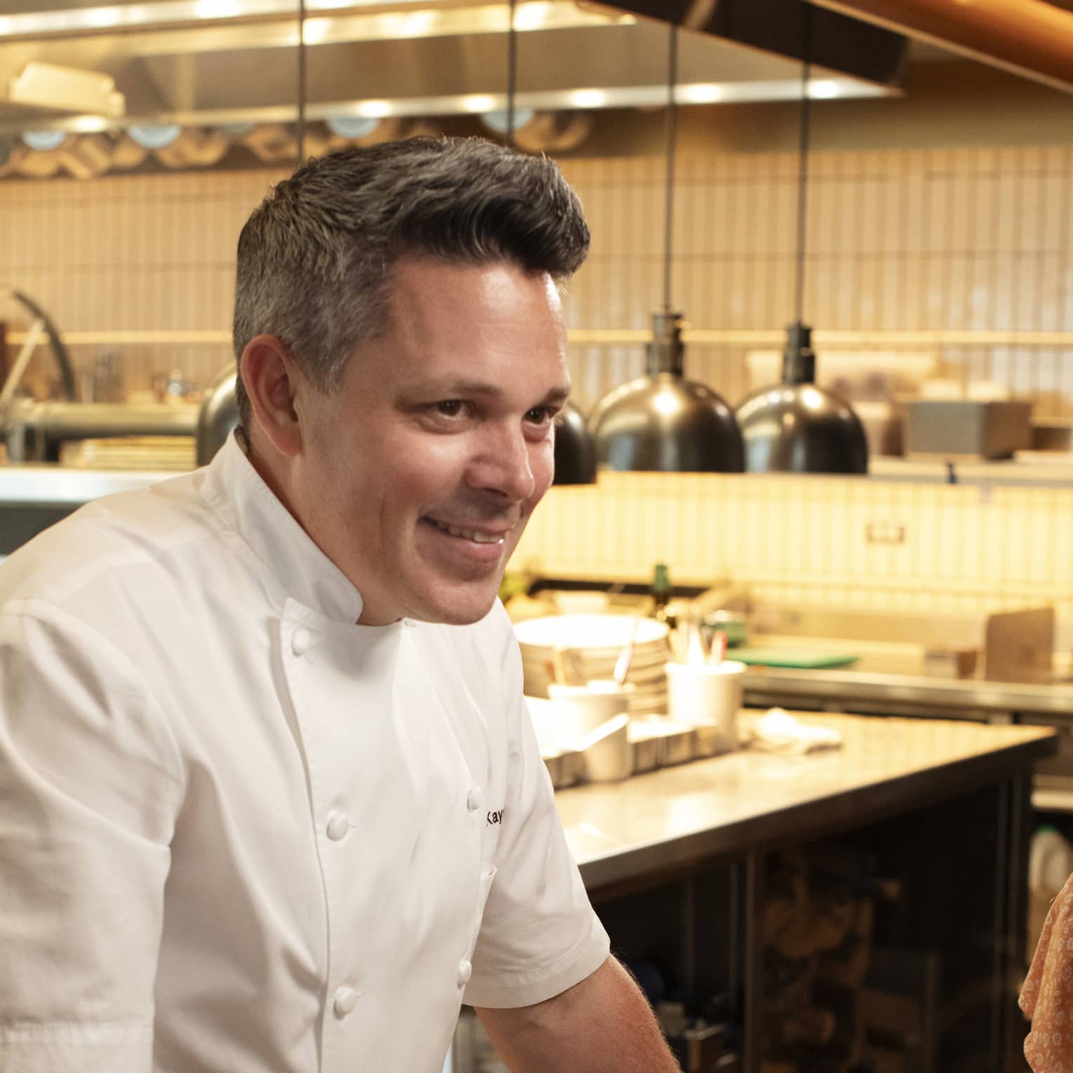 Chef standing beside a dining table in the kitchen talking to guests