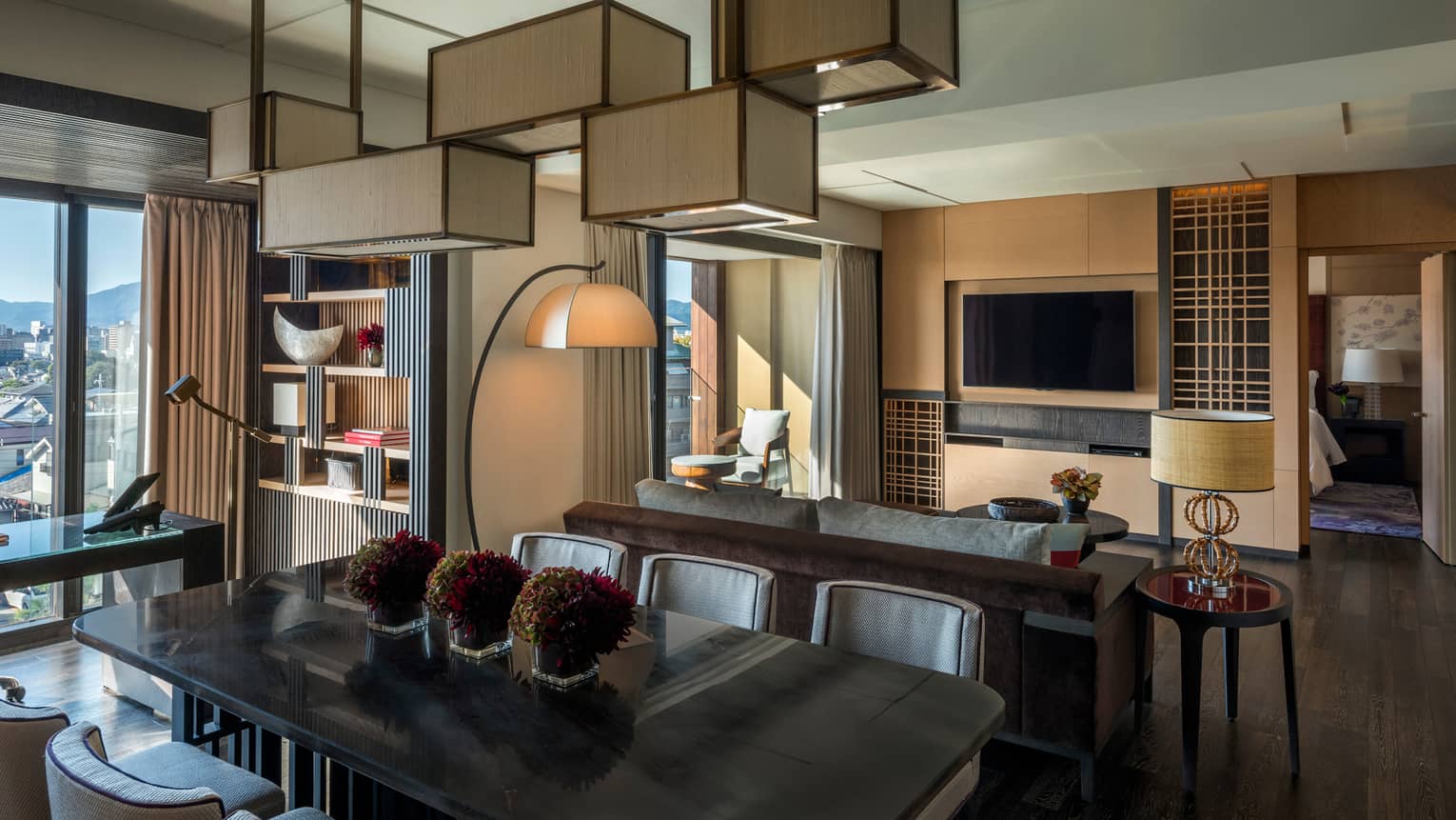 Residential suite with dining table, sofa, wall-mounted TV, floor lamp, bookshelf, desk with leather chair and floor-to-ceiling views of the city skyline
