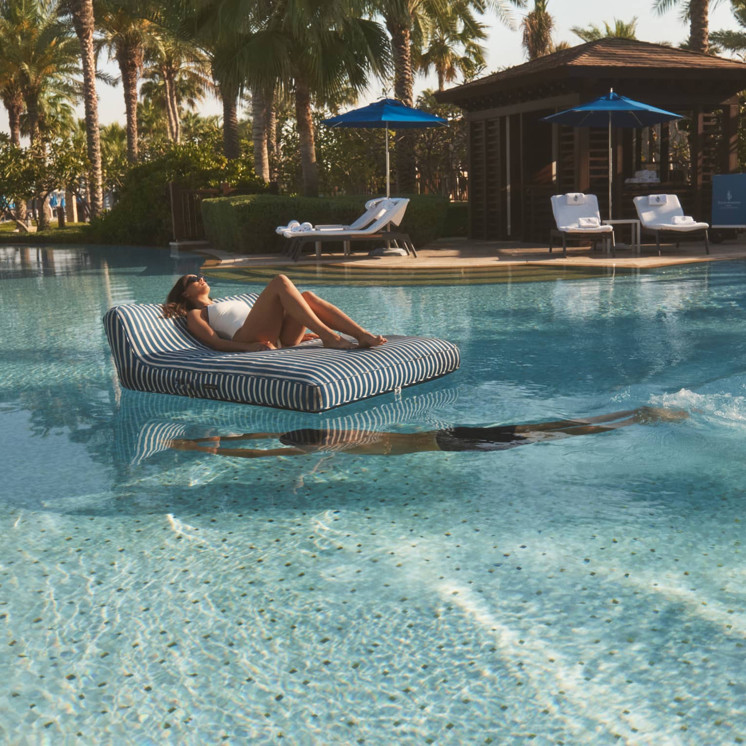 Person lounges on a black-and-white striped floating chair in the middle of a pool as another person swim beneath