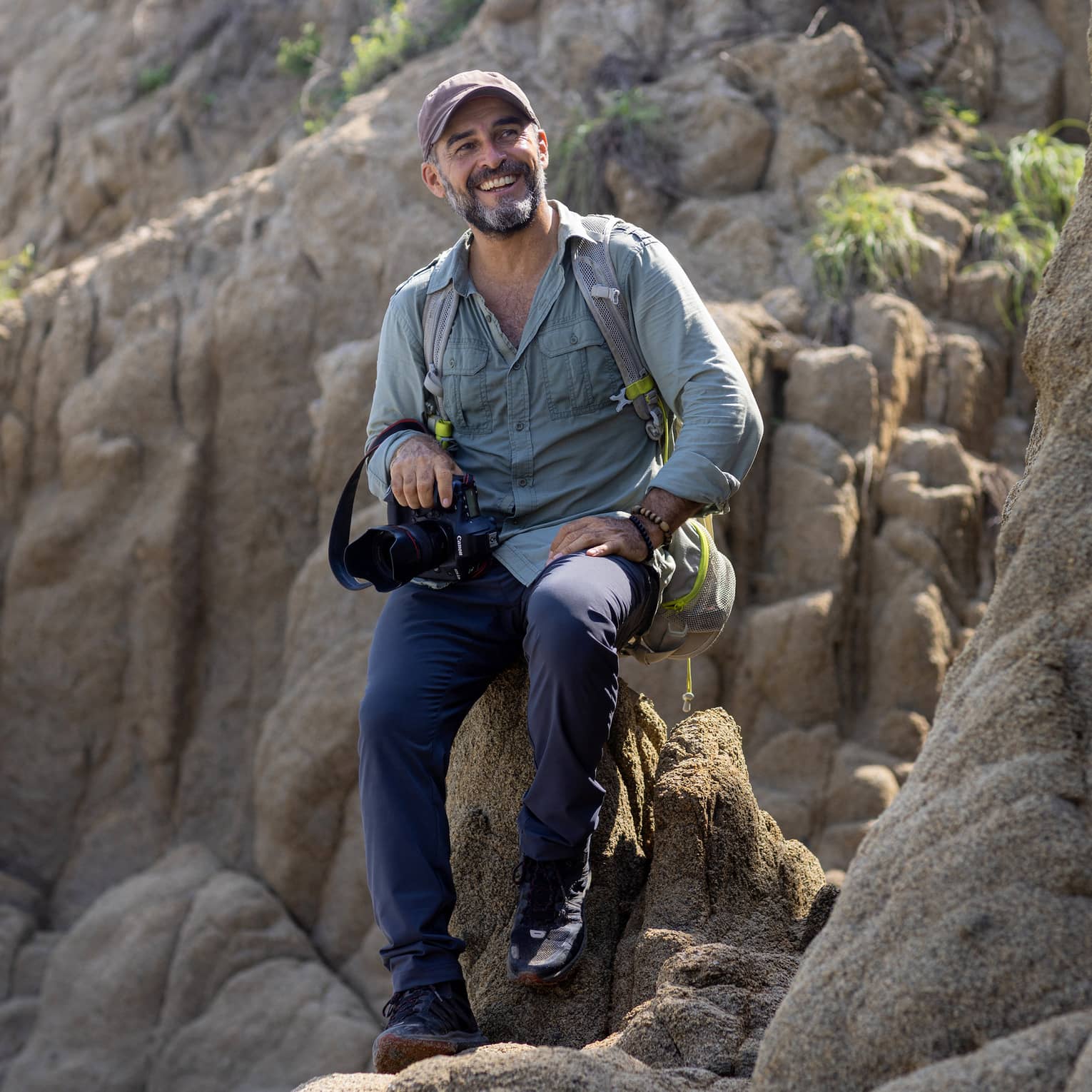 A photographer wearing a backpack and holding a camera in one hand sits atop a rock formation, smiling into the distance.