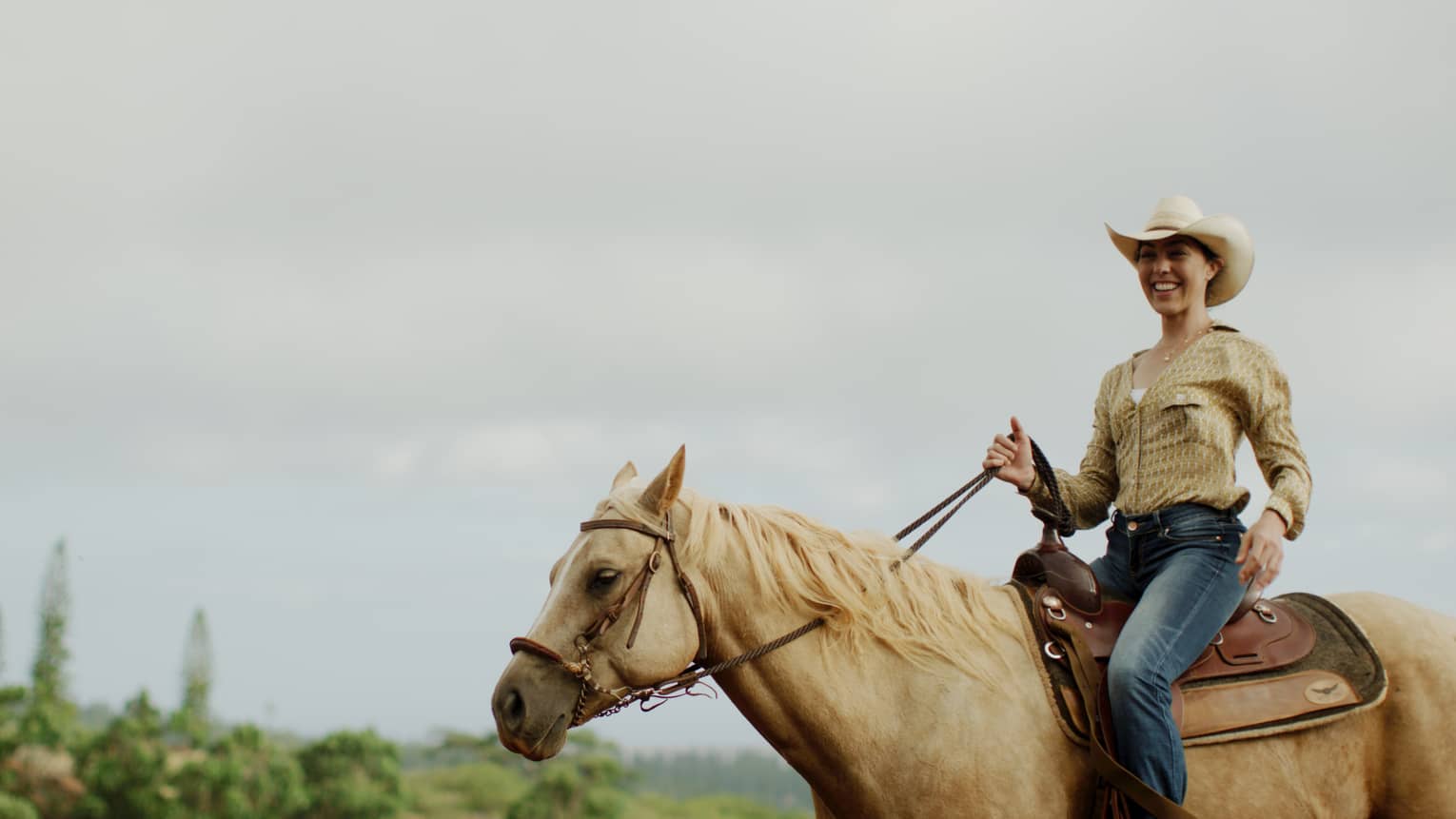 A smiling guest wearing a cowboy hat holds the reins while riding a tan horse under cloudy skies, forest in the distance.
