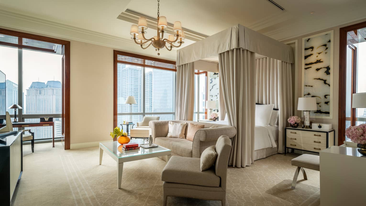 Hotel room canopy bed with white curtains behind sofa and seating area, large windows